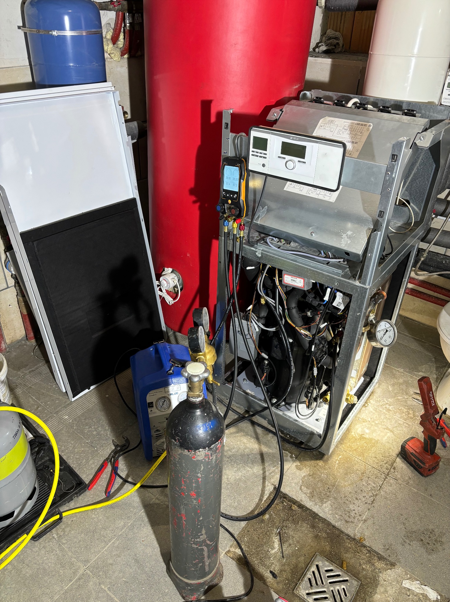 Otwarta obudowa pieca gazowego z podłączonymi przewodami pomiarowymi, butla z gazem obok, widoczne narzędzia, w tle zbiorniki na wodę.