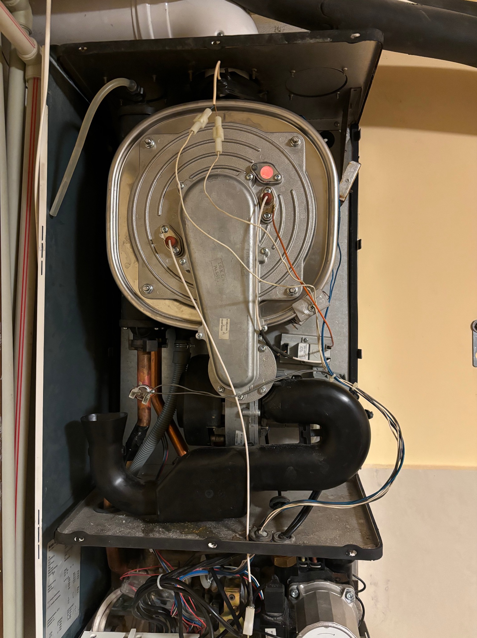 Otwarty piec gazowy z widocznymi elementami wewnętrznymi, w tym srebrną obudową, czarnymi rurami i licznymi przewodami, na tle beżowej ściany.