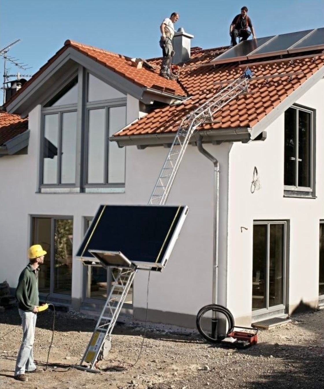 Instalacja paneli słonecznych na dachu domu jednorodzinnego z użyciem specjalistycznej windy dekarskiej; pracownicy w kaskach na dachu, operator windy w żółtym kasku na ziemi.