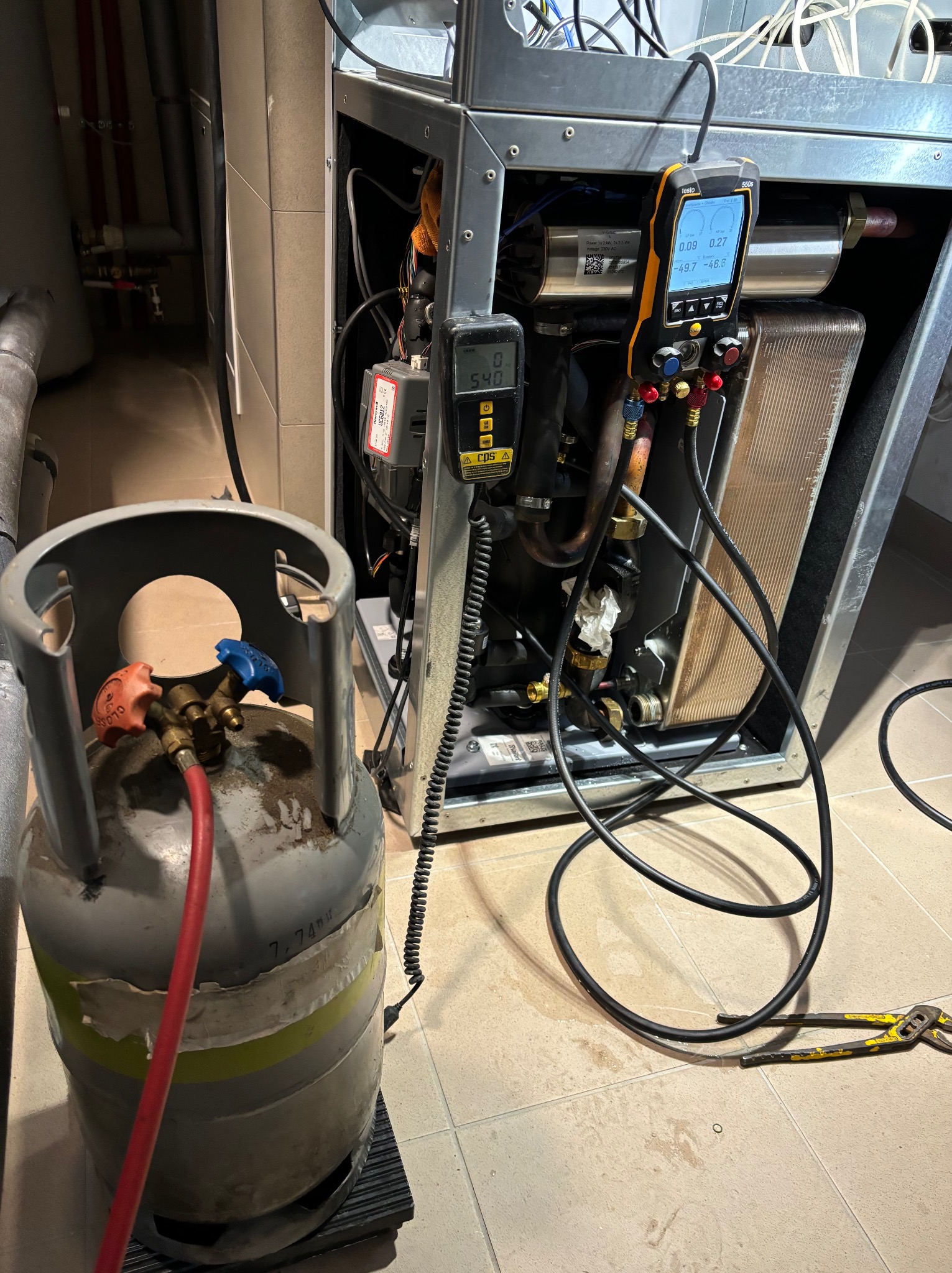 Podłączony cylinder z czynnikiem chłodniczym do urządzenia chłodniczego z widocznymi manometrami cyfrowymi wskazującymi ciśnienie, w tle rury instalacji i płytki podłogowe.