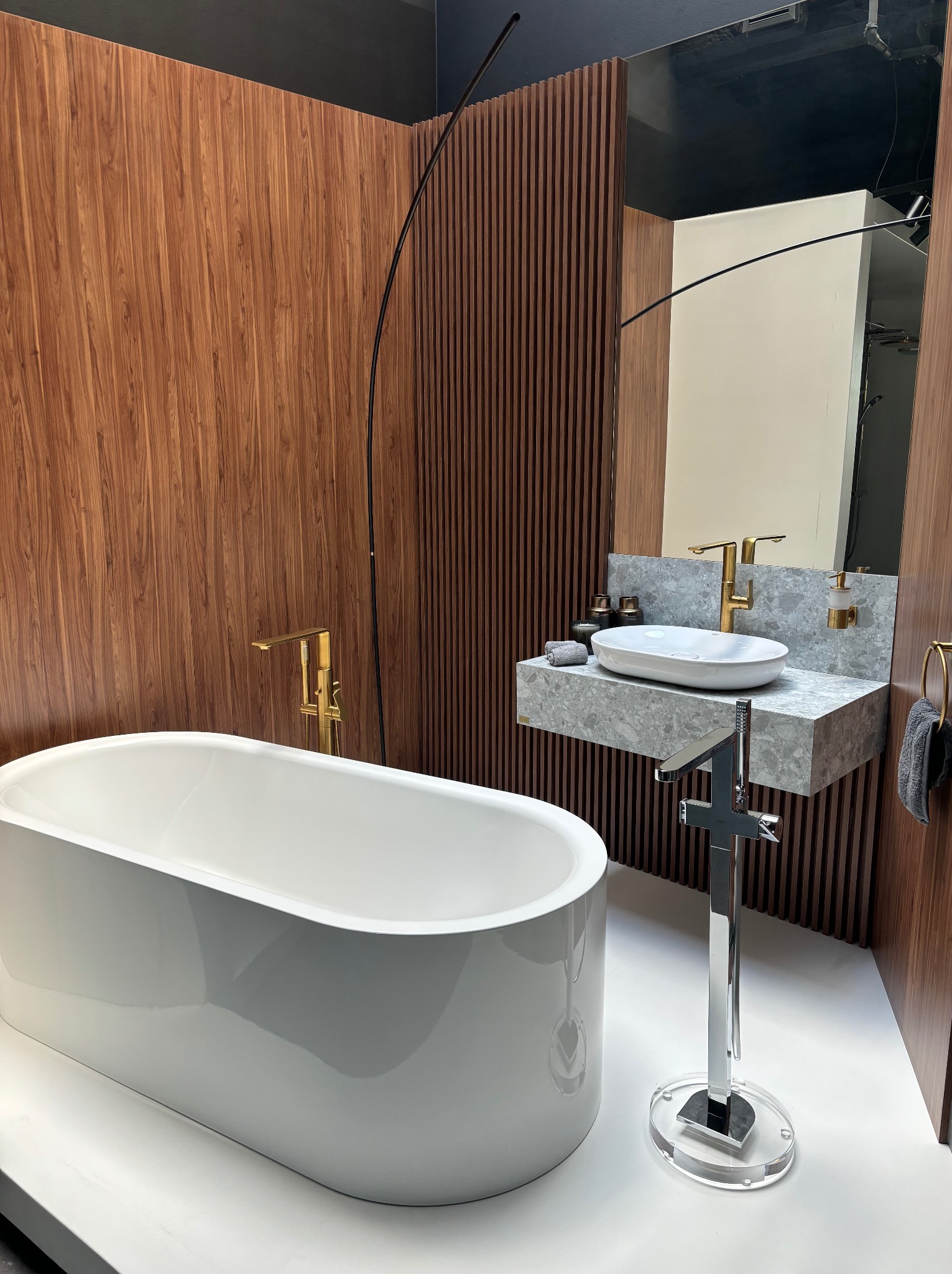 Nowoczesna łazienka w Białymstoku z wolnostojącą białą wanną i umywalką na blacie z szarego kamienia, złota armatura, drewniane panele ścienne.