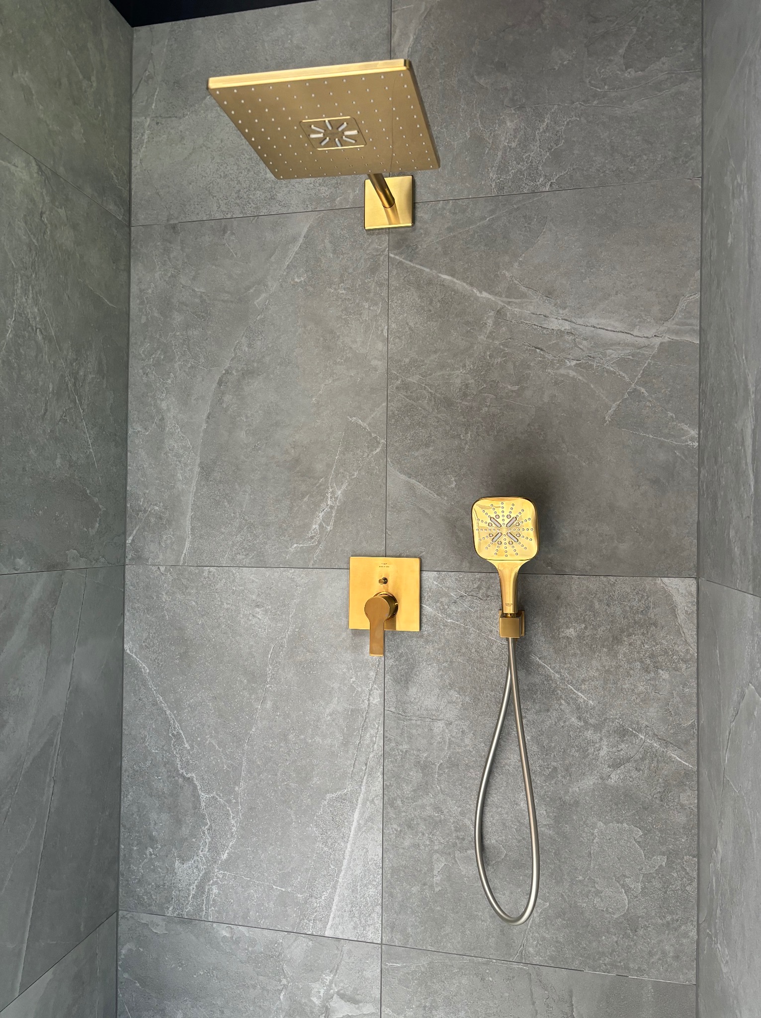 Nowoczesny zestaw prysznicowy w kolorze złotym, z deszczownicą kwadratową i słuchawką, zamontowany na szarych płytkach imitujących marmur.
