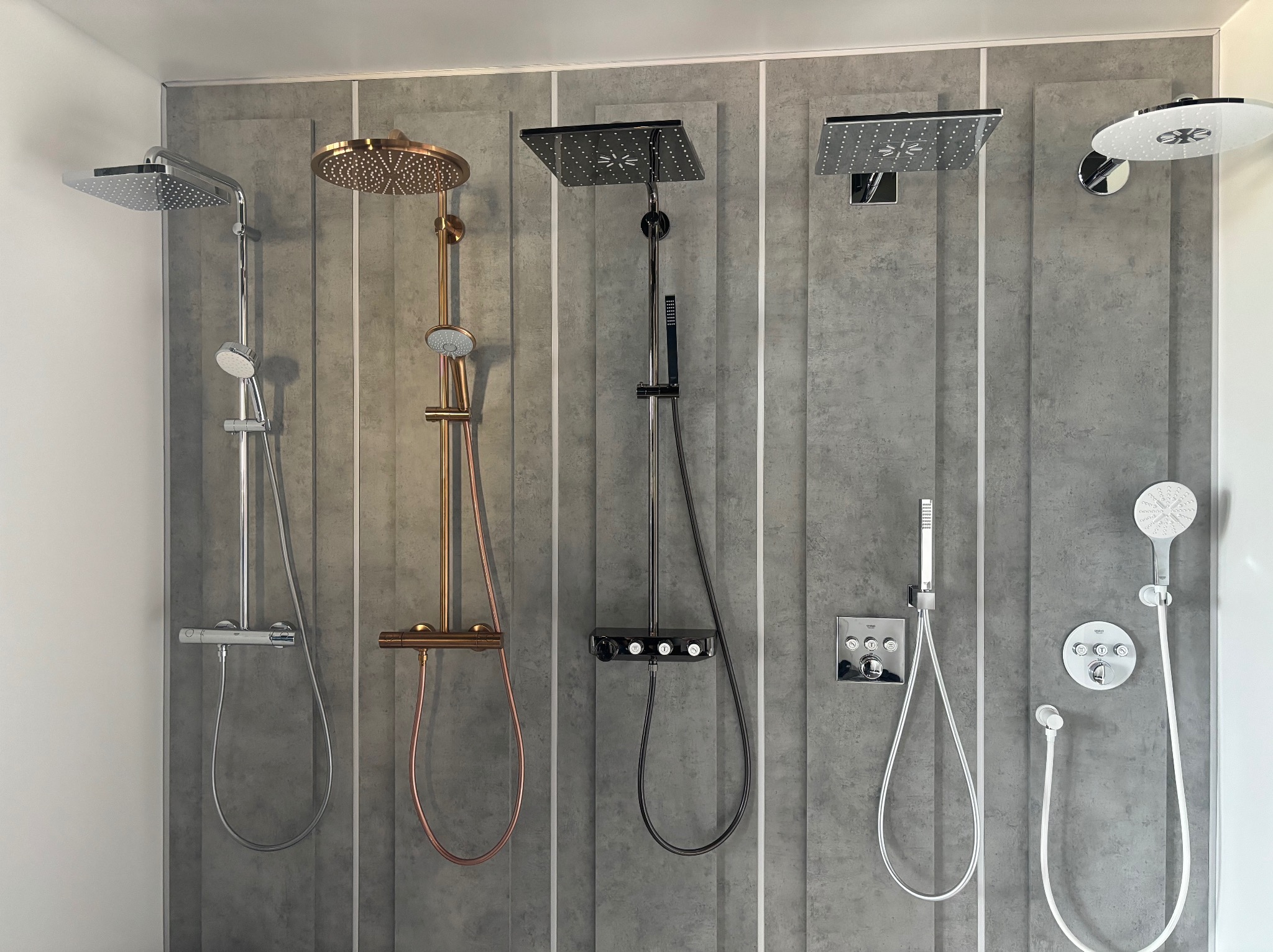 Ekspozycja pięciu różnych zestawów prysznicowych: chromowany kwadratowy, miedziany okrągły, czarny kwadratowy, chromowany kwadratowy z panelem sterującym i biały okrągły, zamontowanych na szarej...