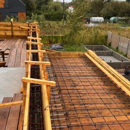 MPJM Michał Mielcarek - Zbrojenie płyty fundamentowej z drewnianym szalunkiem na podkładzie z desek, widok z góry, w tle dom w nowoczesnym stylu i zieleń.