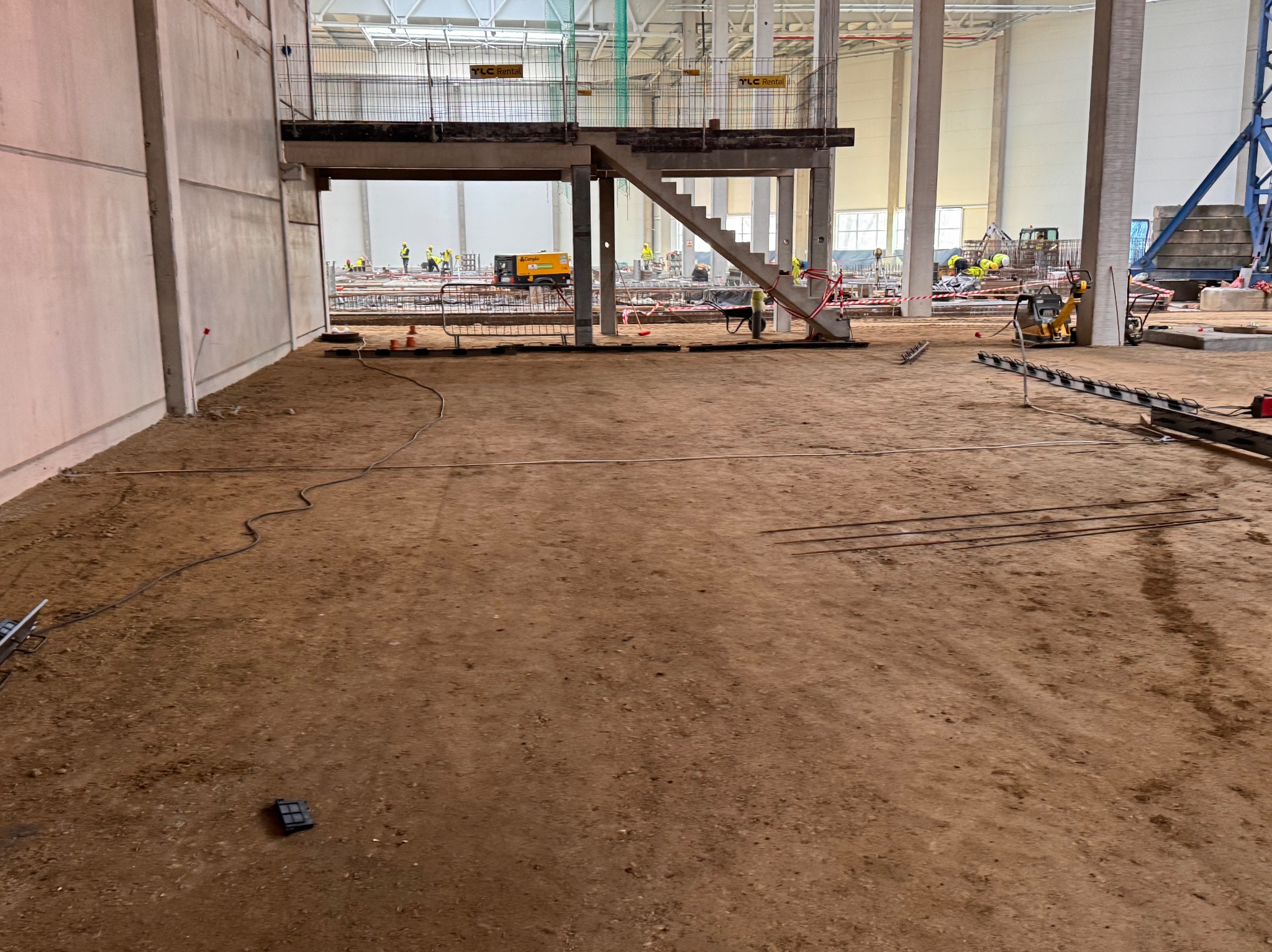 Wnętrze hali w trakcie budowy: widoczna ziemia, betonowe filary, schody z podestem, pracownicy w żółtych kamizelkach oraz sprzęt budowlany w tle, w tym zagęszczarka wibracyjna.