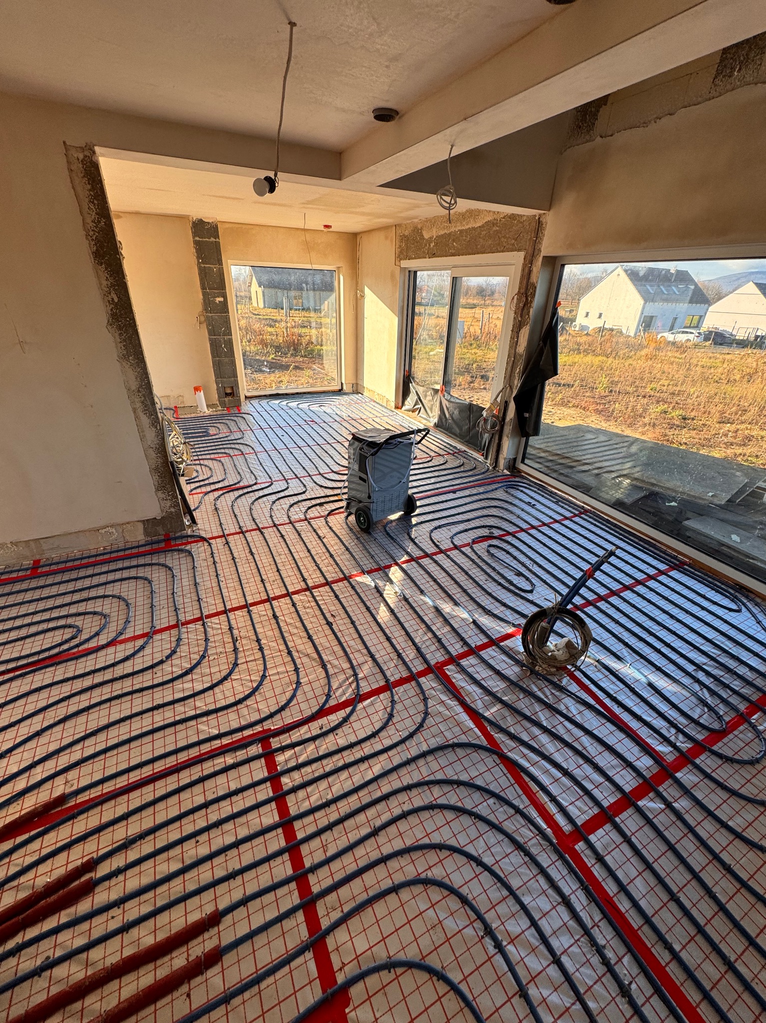 Montaż ogrzewania podłogowego w surowym wnętrzu domu jednorodzinnego, widoczne rury grzewcze układane na folii z siatką, z osuszaczem powietrza w tle.