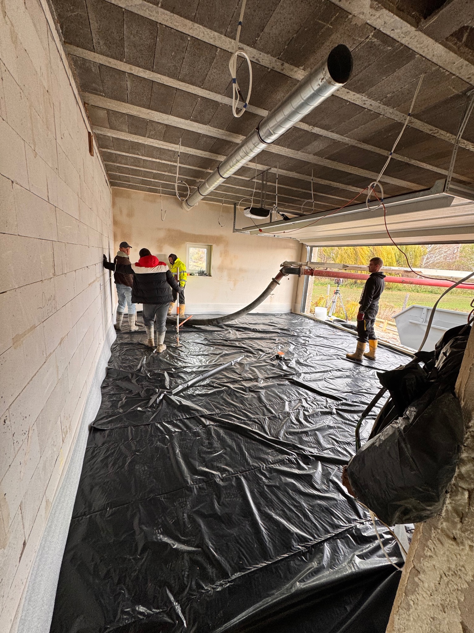 Wnętrze w stanie surowym: robotnicy przygotowują podłoże pod wylewkę, rozkładając czarną folię ochronną na izolacji podłogowej. Widoczna rura doprowadzająca materiał do wylewki oraz ściany z betonu...