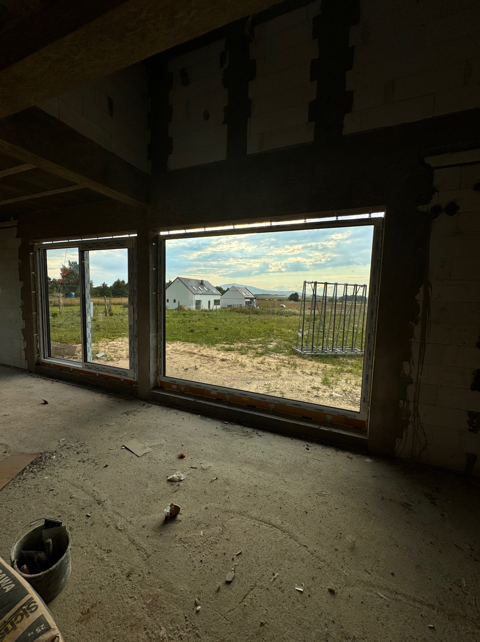 Surowe wnętrze domu w trakcie budowy, widoczne okna z widokiem na krajobraz wiejski i drugi dom, betonowa podłoga z resztkami materiałów budowlanych, ściany z bloczków.