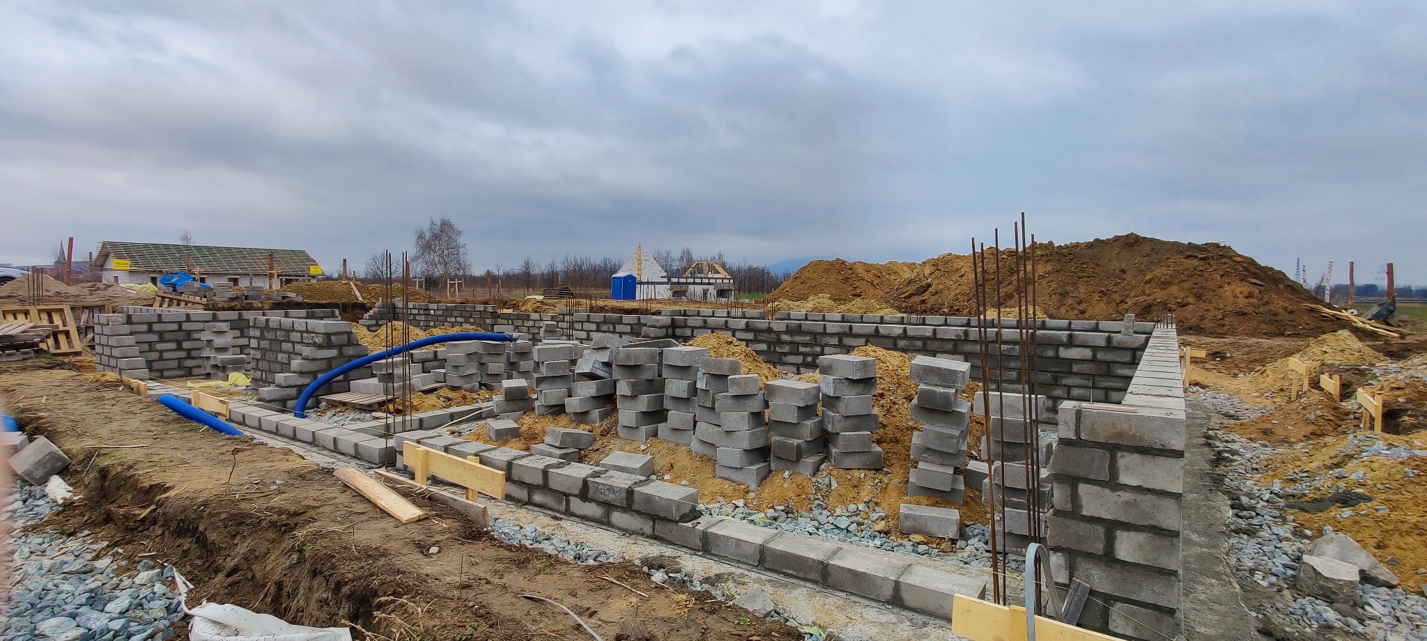 Fundamenty domu w trakcie budowy: ściany z bloczków betonowych, wystające pręty zbrojeniowe, niebieski wąż drenażowy, ziemia i gruz, w tle budynki pod konstrukcją dachu