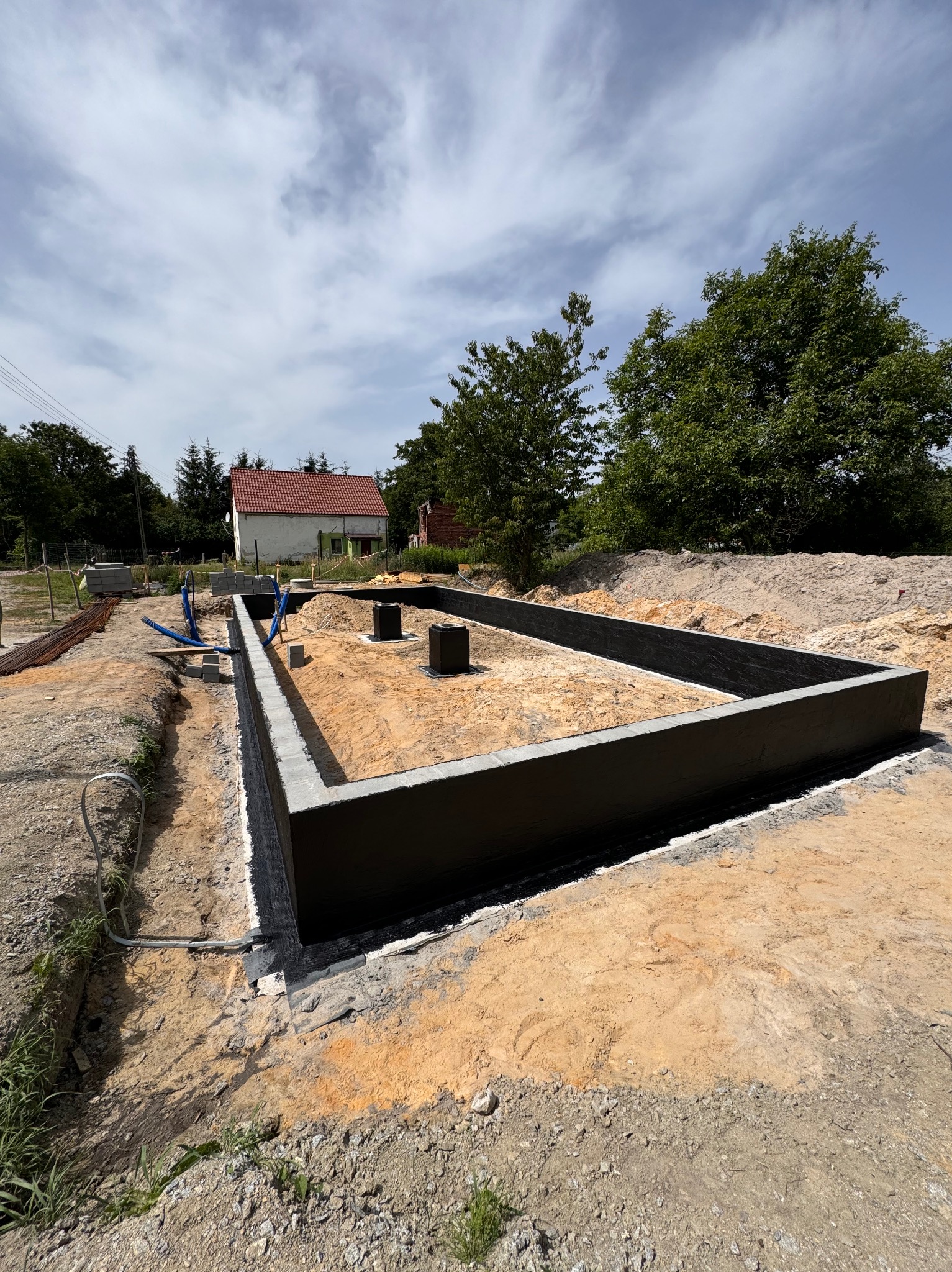 Zalane fundamenty budynku z izolacją przeciwwilgociową, widoczne szalunki i przygotowany teren pod budowę, w tle dom z czerwoną dachówką i zieleń.