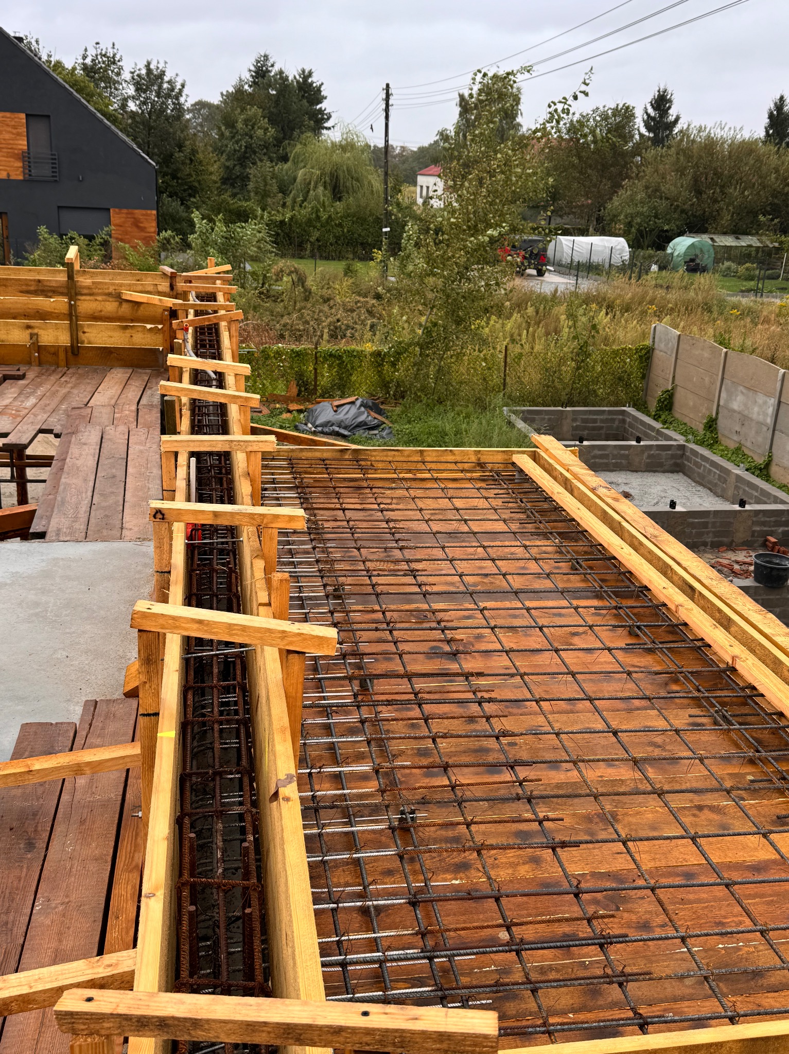 Zbrojenie płyty fundamentowej z drewnianym szalunkiem na podkładzie z desek, widok z góry, w tle dom w nowoczesnym stylu i zieleń.