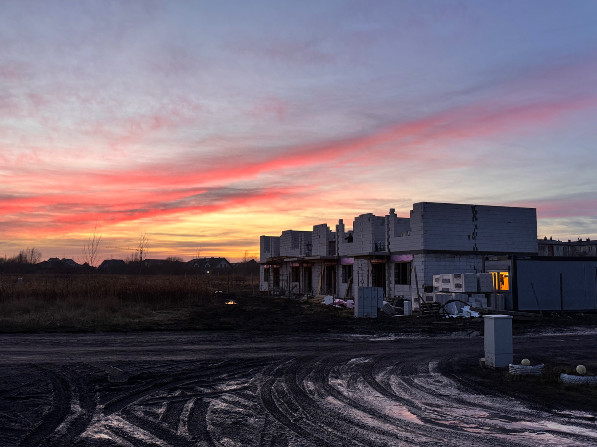 Wczesny etap budowy szeregu domów z bloczków, widoczne ślady opon na błotnistym terenie, zachodzące słońce maluje niebo na czerwono i pomarańczowo, w oknie jednego z domów widać zapalone światło.