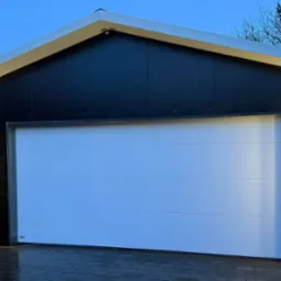 Nowoczesny garaż z segmentową bramą w kolorze białym, czarną elewacją i oświetleniem LED po bokach drzwi wejściowych, sfotografowany o zmierzchu.