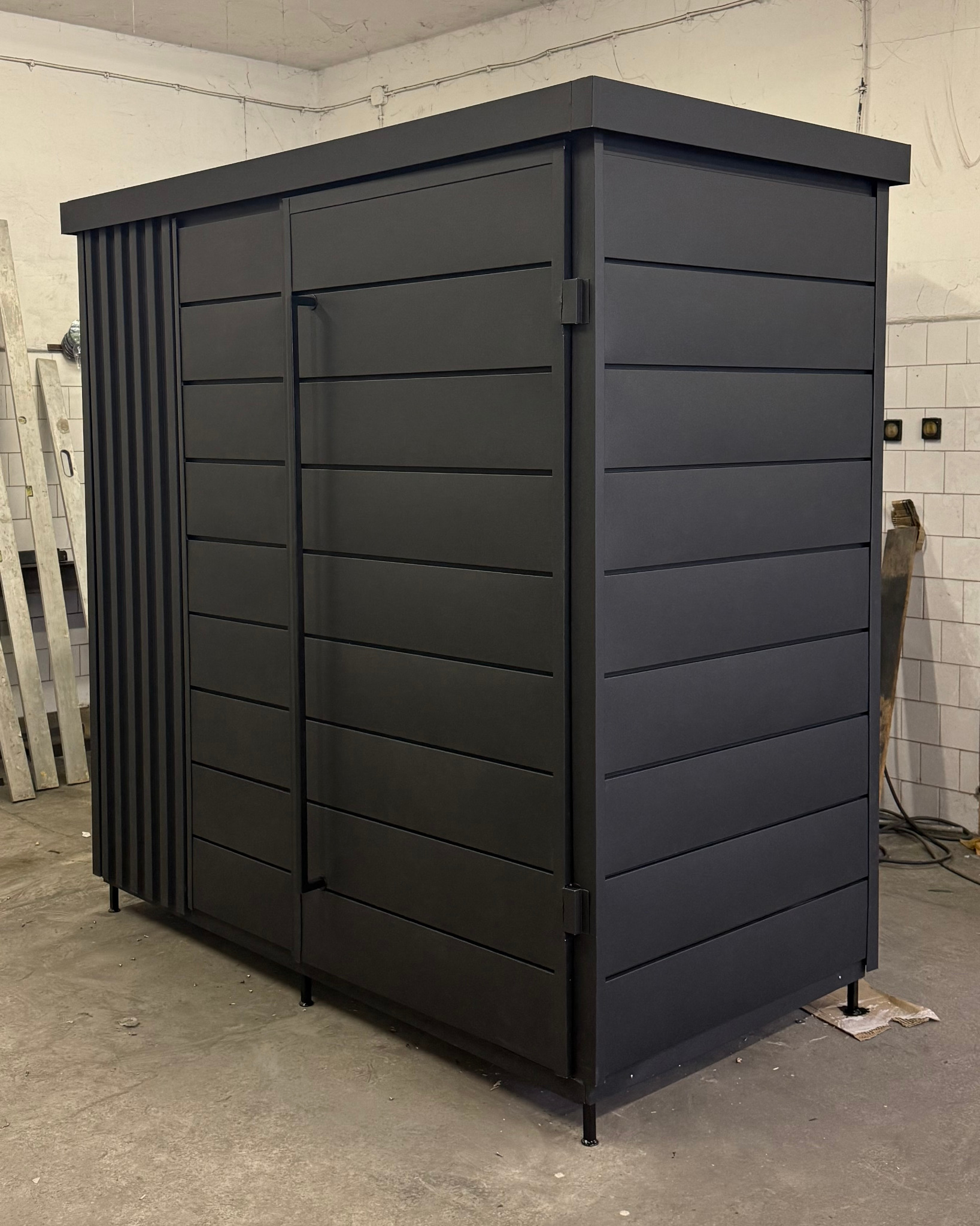 Czarna, metalowa szafa ogrodowa z lamelami po lewej stronie, widoczna w warsztacie.