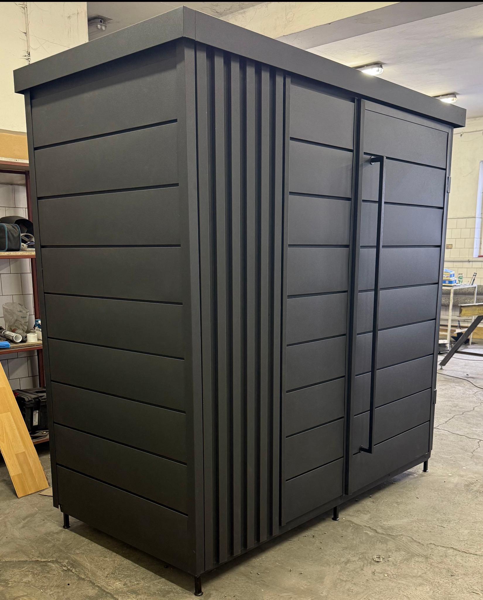 Ciemnoszara metalowa szafa ogrodowa z poziomymi panelami i pionowymi zdobieniami, widoczna w warsztacie, z długimi uchwytami na drzwiach.