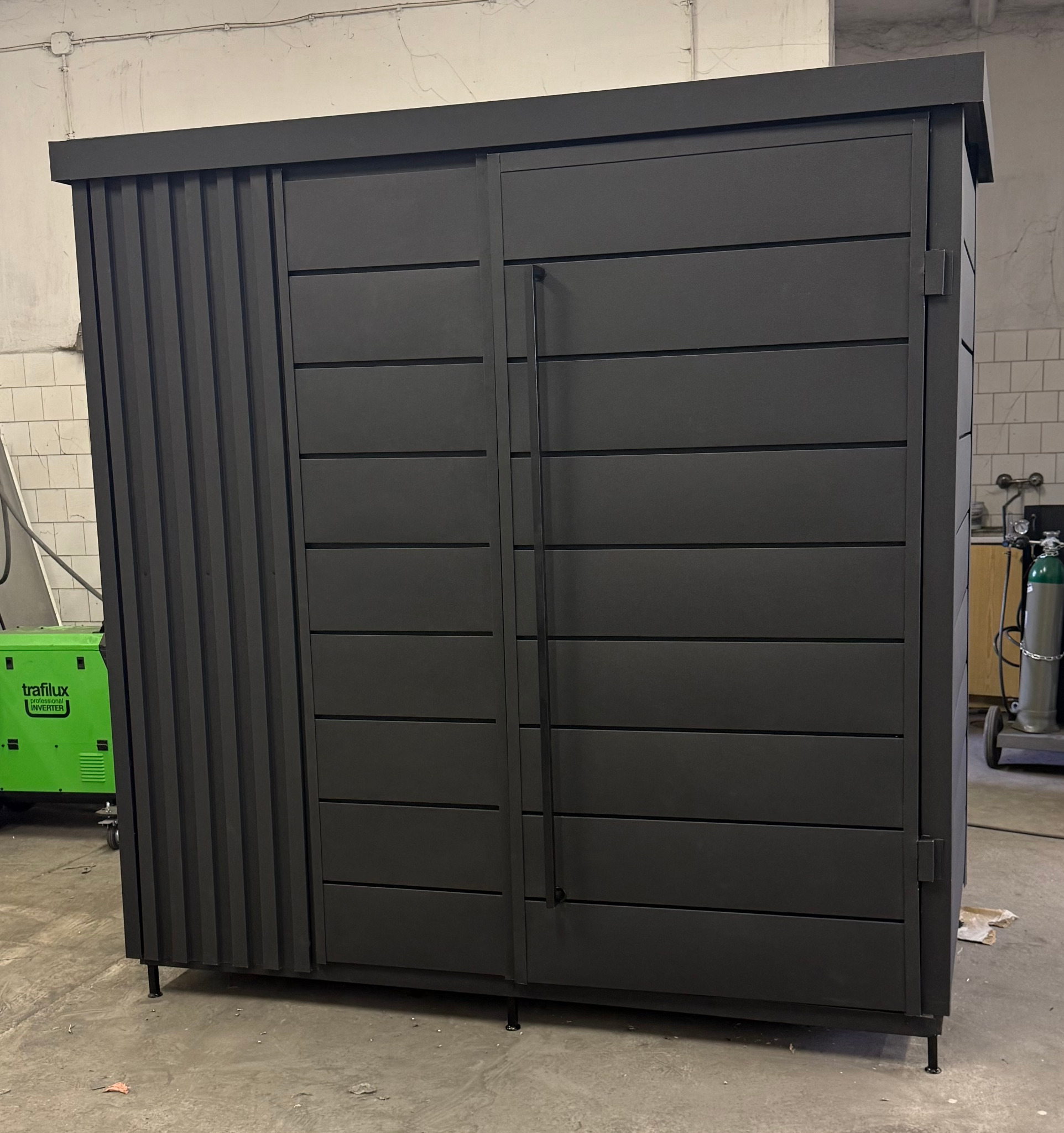 Czarna, metalowa szafa ogrodowa z przesuwnymi drzwiami o horyzontalnym układzie paneli, z widocznym zielonym spawarką inwertorową w tle warsztatu.
