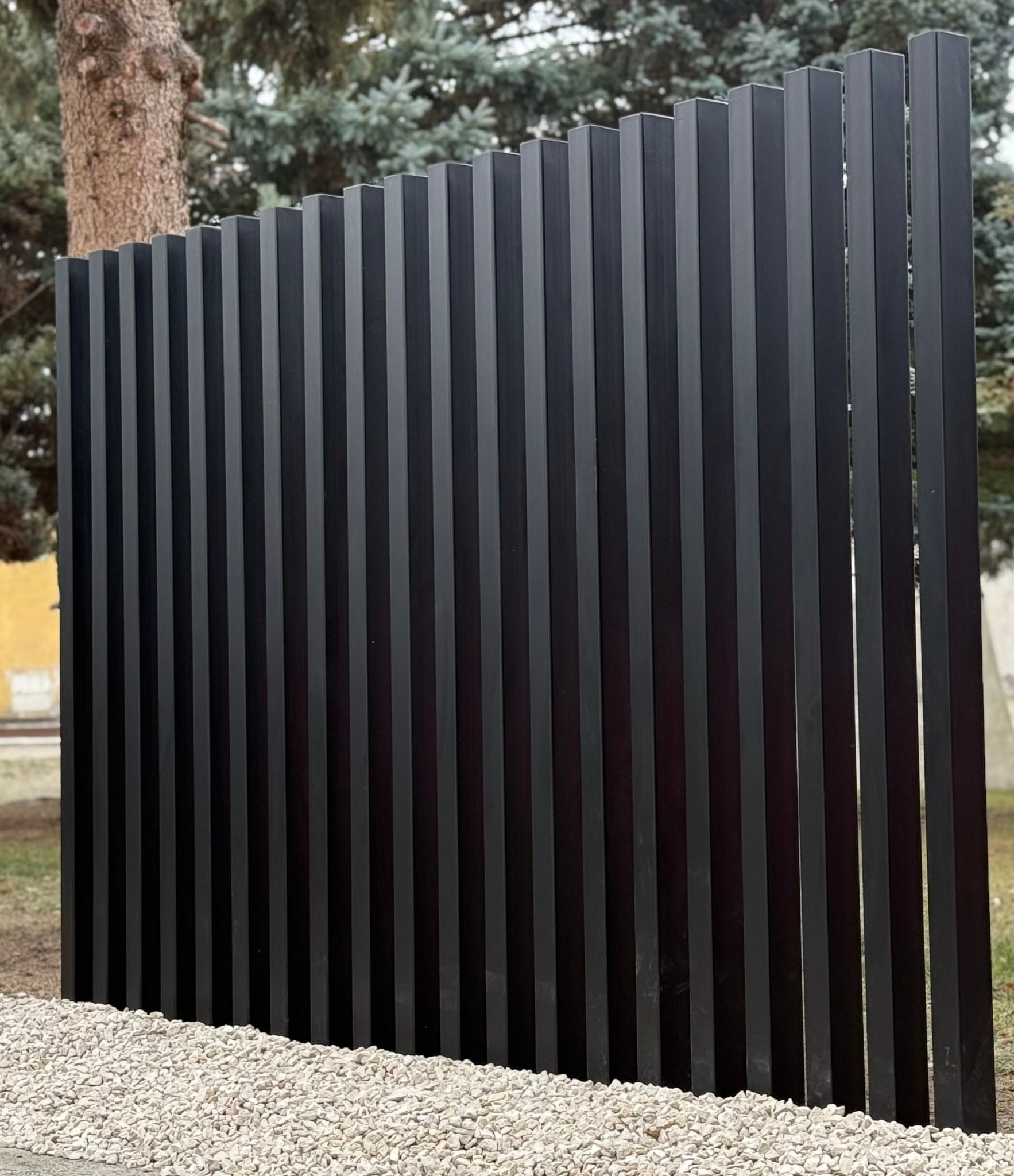Nowoczesne ogrodzenie pionowe z metalowych paneli w kolorze antracytowym, otoczone białym żwirem, zrealizowane w Kamieniu na tle zieleni.