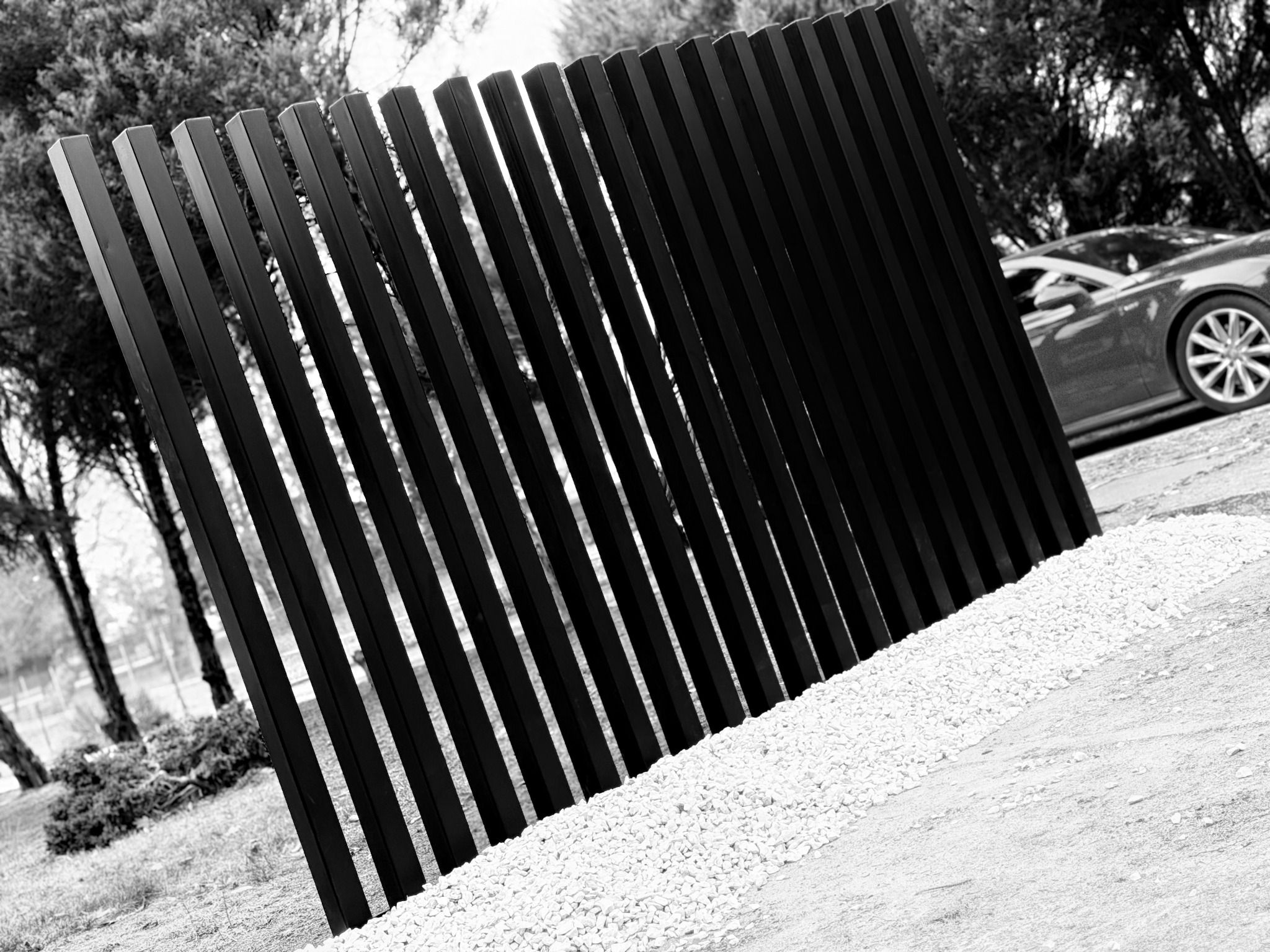 Czarno-białe zdjęcie nowoczesnego ogrodzenia z pionowych, prostokątnych profili, z samochodem w tle i białym żwirem u podstawy.