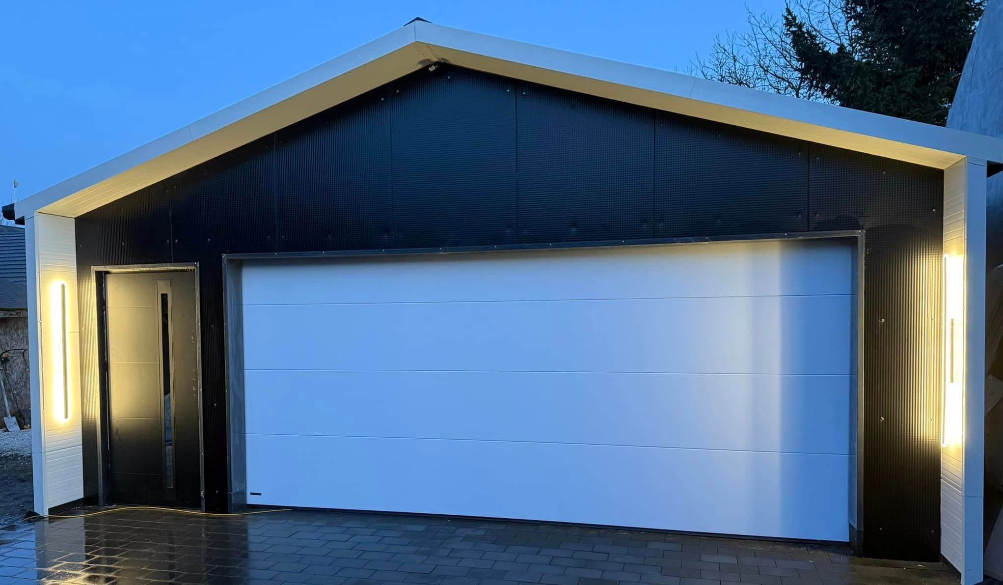 Nowoczesny garaż z segmentową bramą w kolorze białym, czarną elewacją i oświetleniem LED po bokach drzwi wejściowych, sfotografowany o zmierzchu.