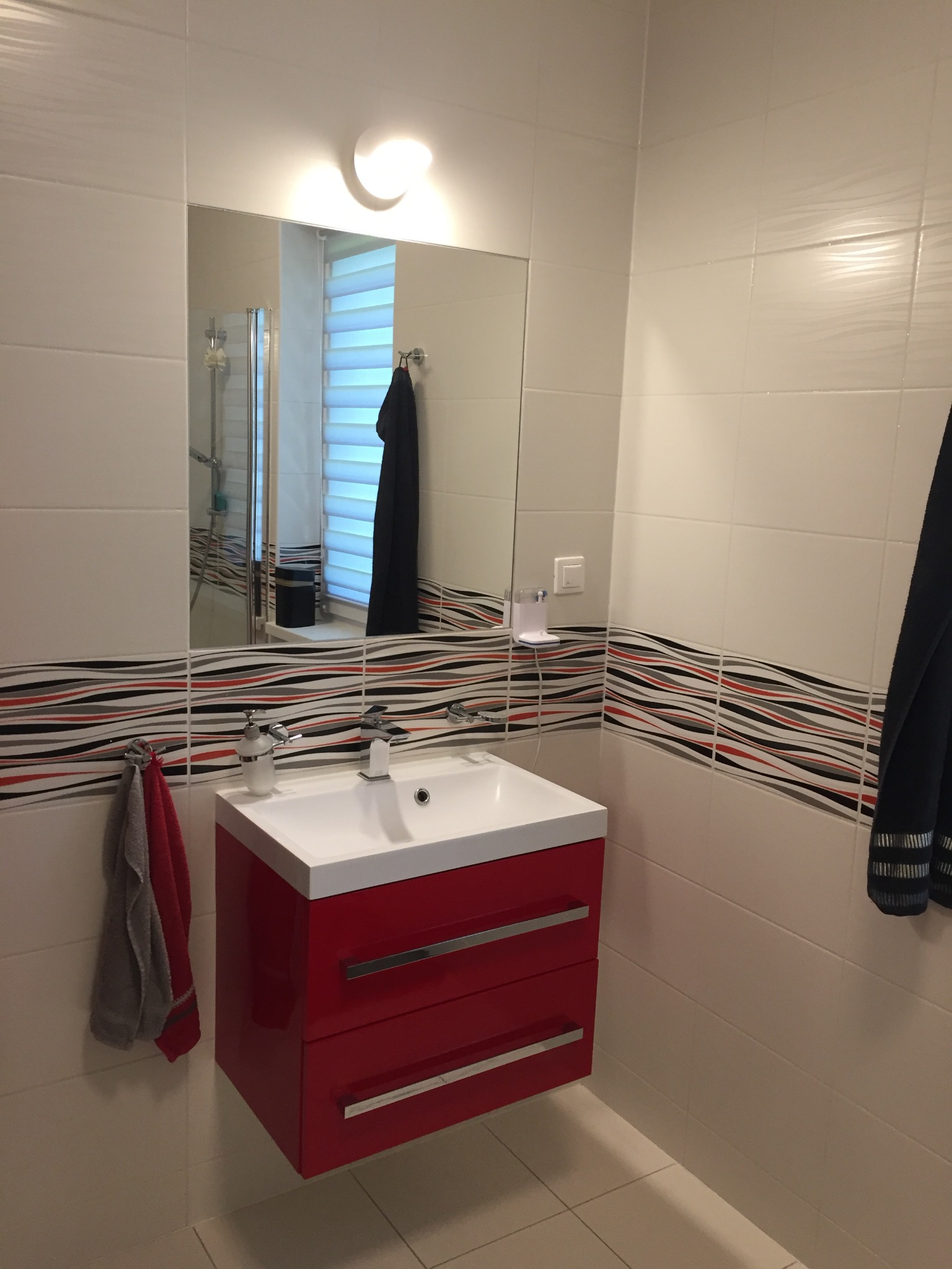Biała łazienka z czerwonymi szafkami pod umywalką, wzorzyste płytki dekoracyjne na ścianie, lustro z widoczną kabiną prysznicową i roletami w oknie.
