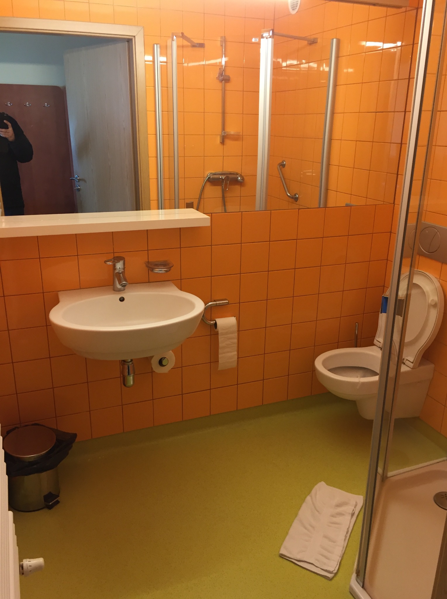 Pomarańczowa łazienka z żółtą podłogą, widoczna umywalka, toaleta i kabina prysznicowa z ręcznikiem na podłodze.