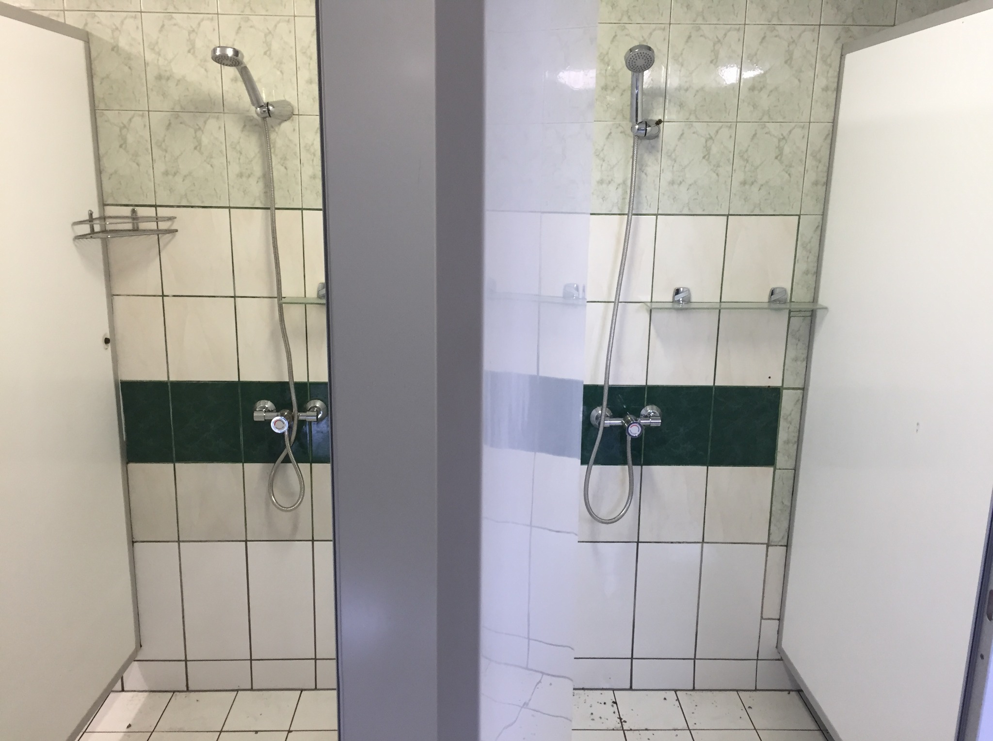 Dwa boksy prysznicowe z białymi drzwiami i płytkami w odcieniach bieli i zieleni, widoczne chromowane słuchawki prysznicowe i półki.