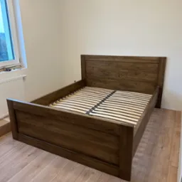 Drewniane łóżko z listwowym stelażem, ustawione na panelach podłogowych, widok z przodu, neutralne oświetlenie.