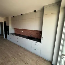 Jasnoszara zabudowa kuchenna z czarnym blatem i baterią, ceglana ściana nad blatem, oświetlenie punktowe sufitowe, drewniana podłoga.