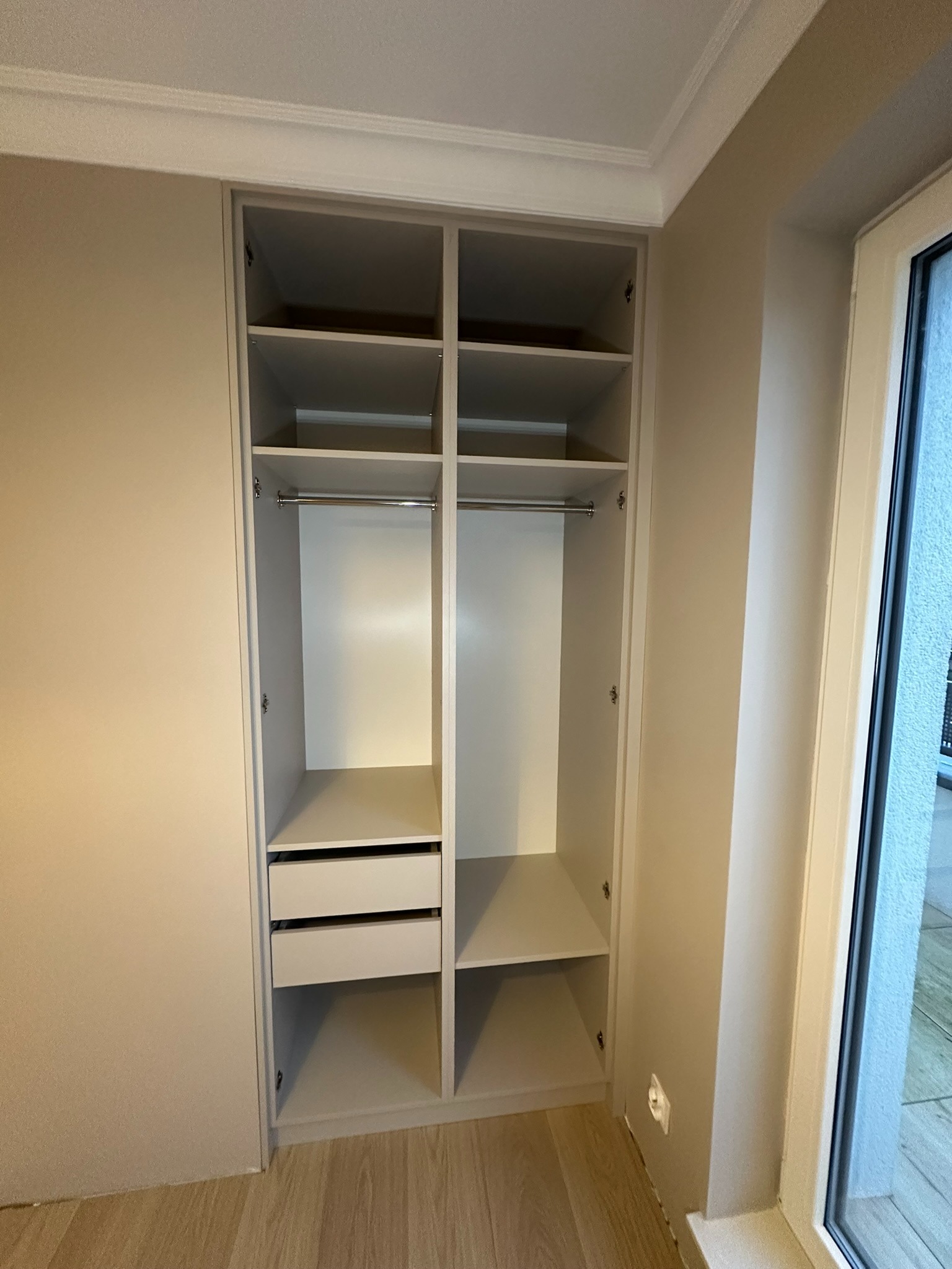 Jasnoszara szafa wnękowa z półkami, drążkiem i dwiema szufladami, wbudowana między ścianę a okno, widok z przodu.