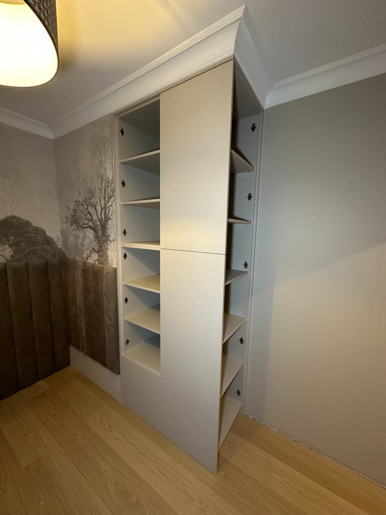 Biała szafa wnękowa z otwartymi półkami, dopasowana do listwy sufitowej, na tle ściany z tapetą z motywem drzewa, widoczna podłoga z paneli.