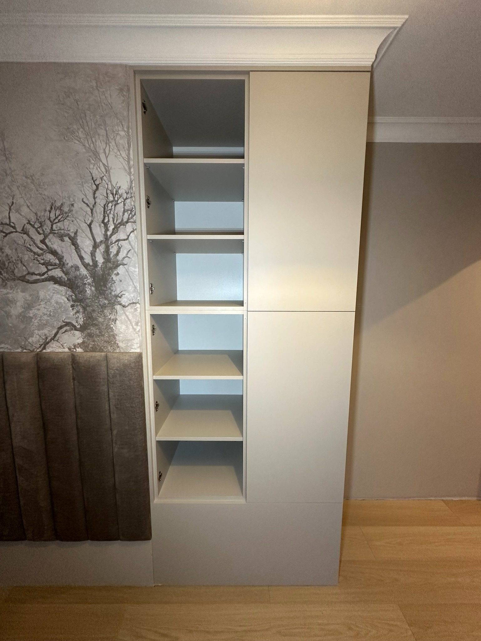 Biała szafa wnękowa z otwartymi półkami i zamkniętymi drzwiami, dopasowana do zagłówka łóżka i tapety z motywem drzewa w sypialni.