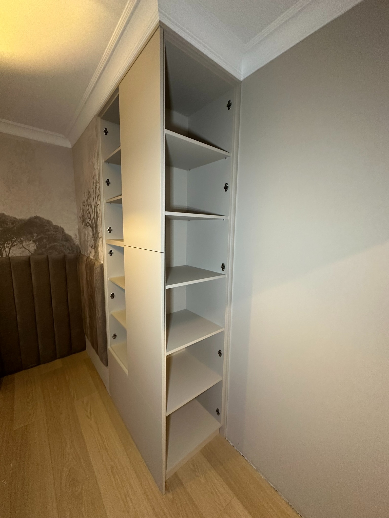 Jasnoszara szafa wnękowa z otwartymi półkami i zamkniętą górną częścią, dopasowana do wnętrza sypialni z tapetą w motywy drzew i panelem tapicerowanym przy łóżku.