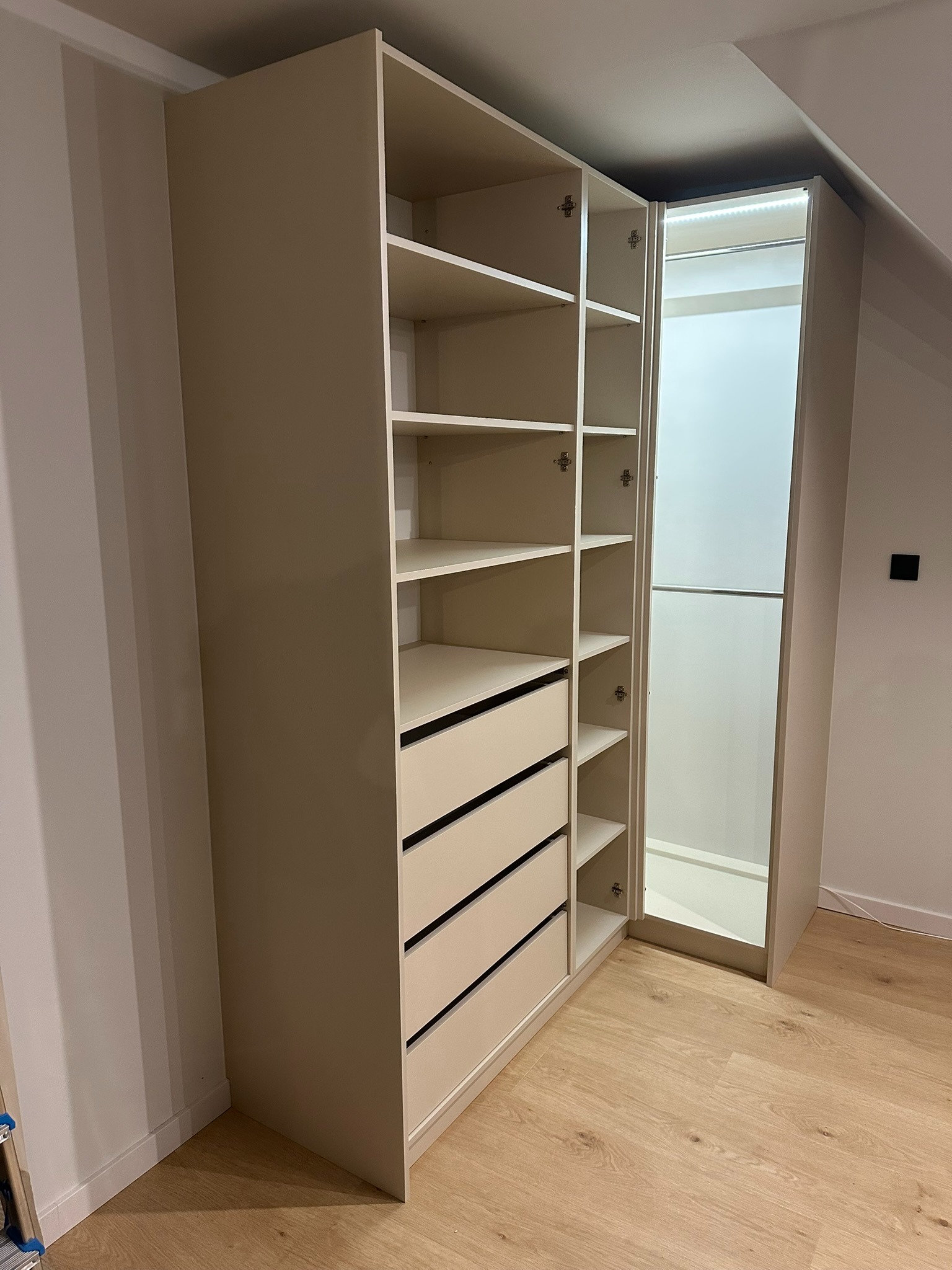 Beżowa szafa wnękowa z półkami i szufladami, oświetlenie LED wewnątrz, widok z boku na jasnej podłodze, zamontowana w skosie poddasza.