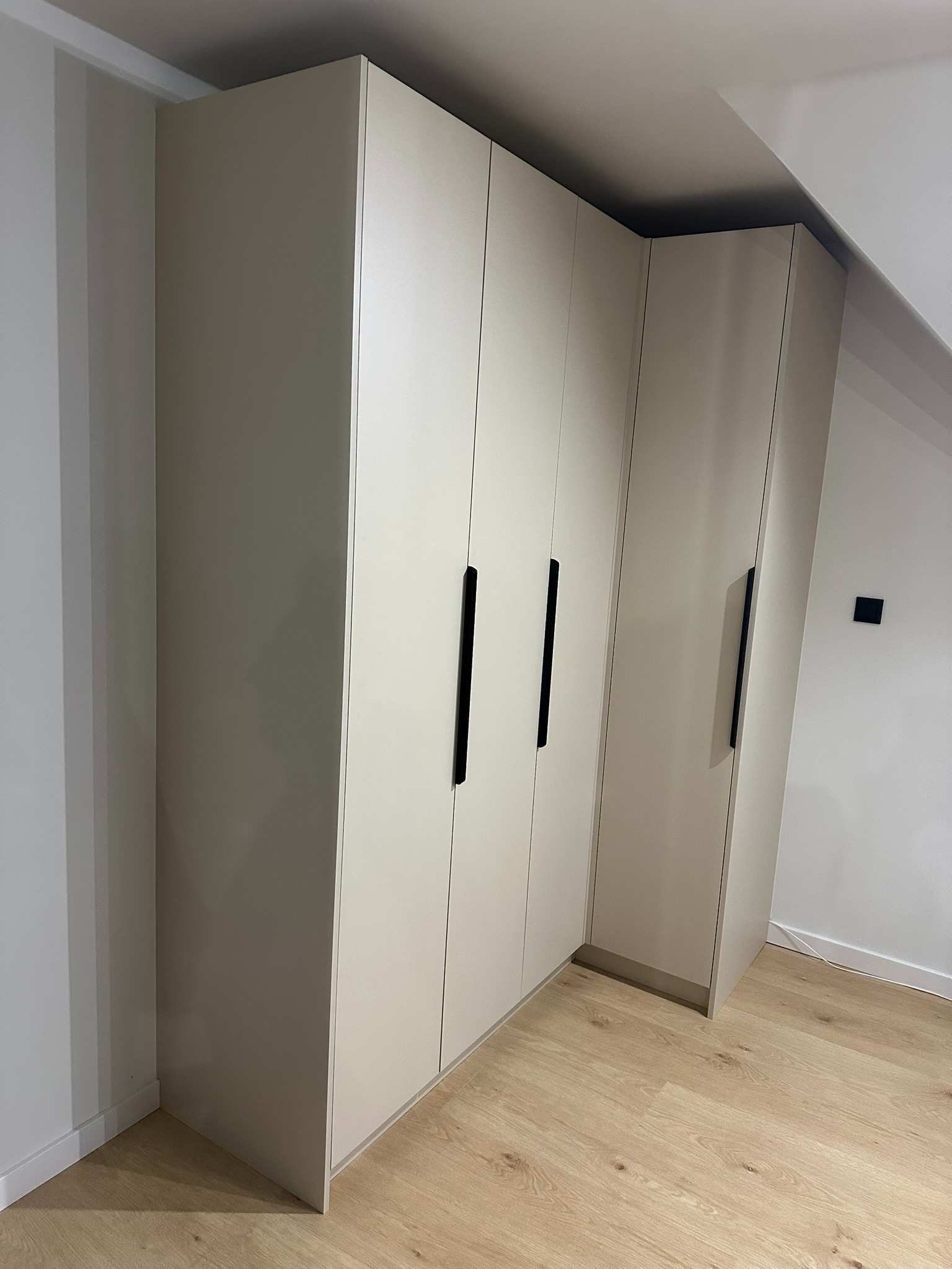 Jasna, zabudowana szafa wnękowa z długimi, czarnymi uchwytami, dopasowana do skosu dachu, na tle jasnych ścian i drewnianej podłogi.