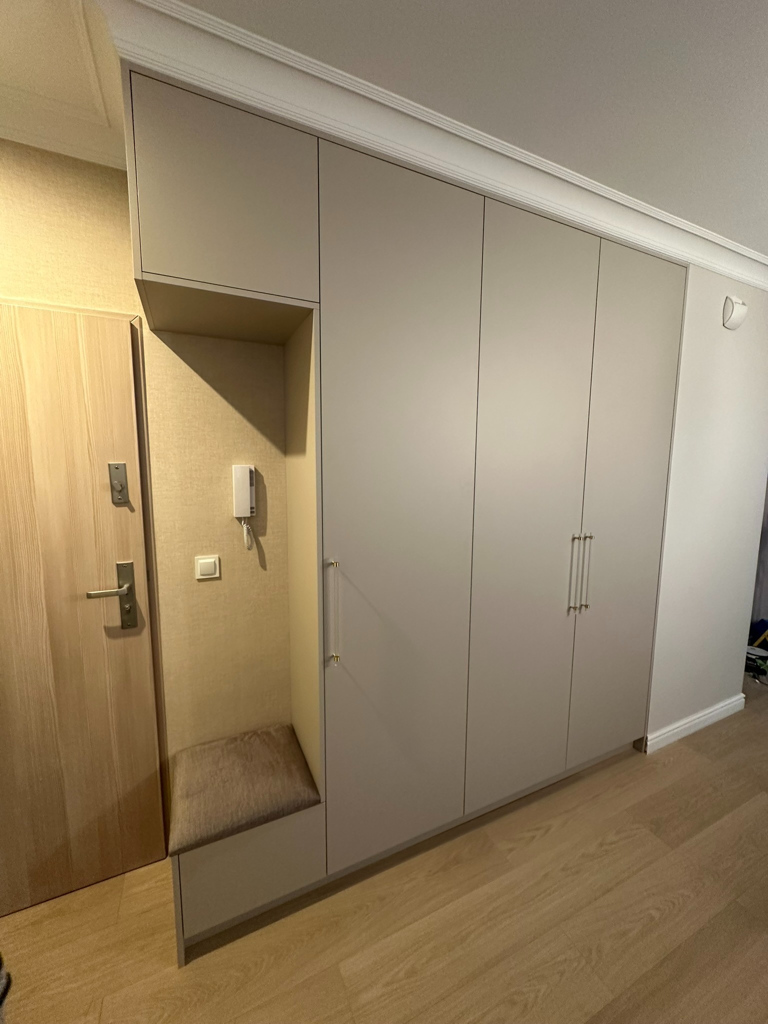 Jasnoszara szafa wnękowa z siedziskiem i wieszakiem, dopasowana do wysokości pomieszczenia, z długimi, przezroczystymi uchwytami, obok drzwi wejściowych w mieszkaniu.