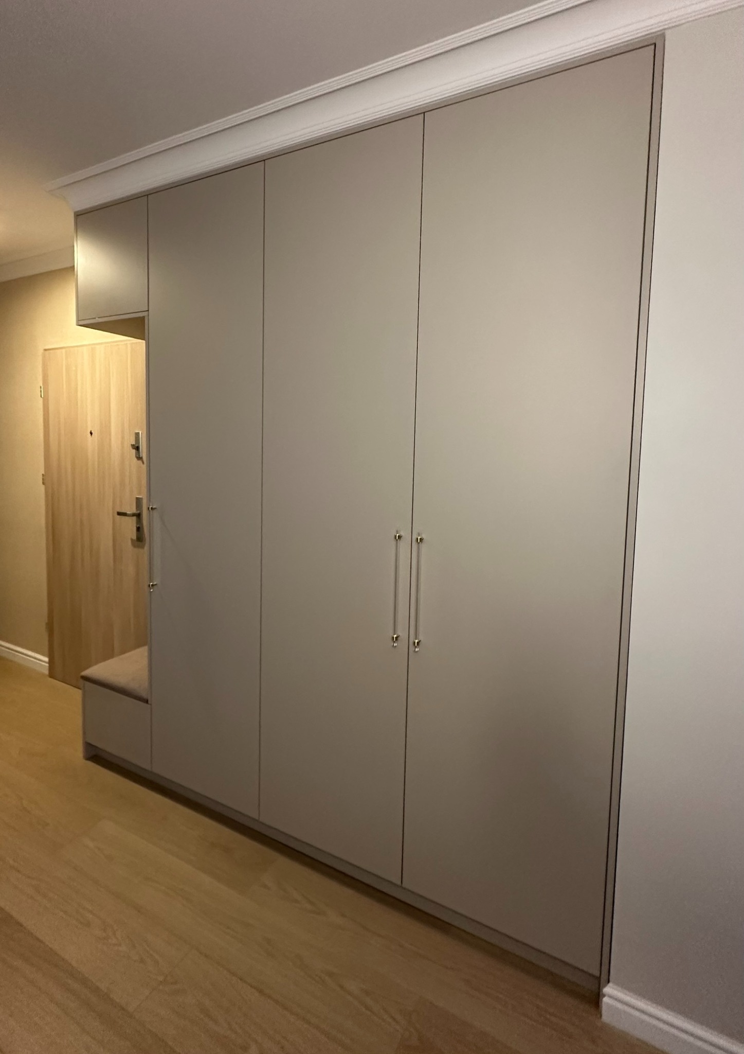Jasnoszara szafa wnękowa z siedziskiem przy wejściu, minimalistyczny design, drewniana podłoga, srebrne uchwyty, listwa dekoracyjna pod sufitem.