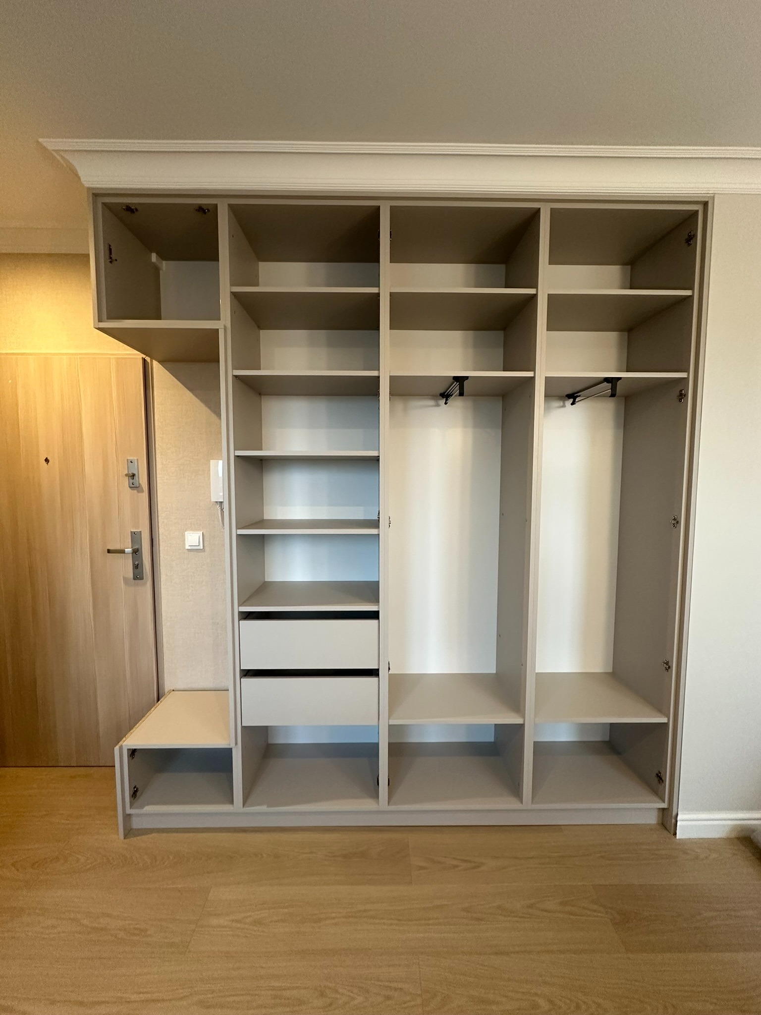 Jasnoszara szafa wnękowa z półkami, szufladami i drążkami na ubrania, dopasowana do wysokości pomieszczenia, z dekoracyjną listwą na górze, widoczna obok drzwi wejściowych.