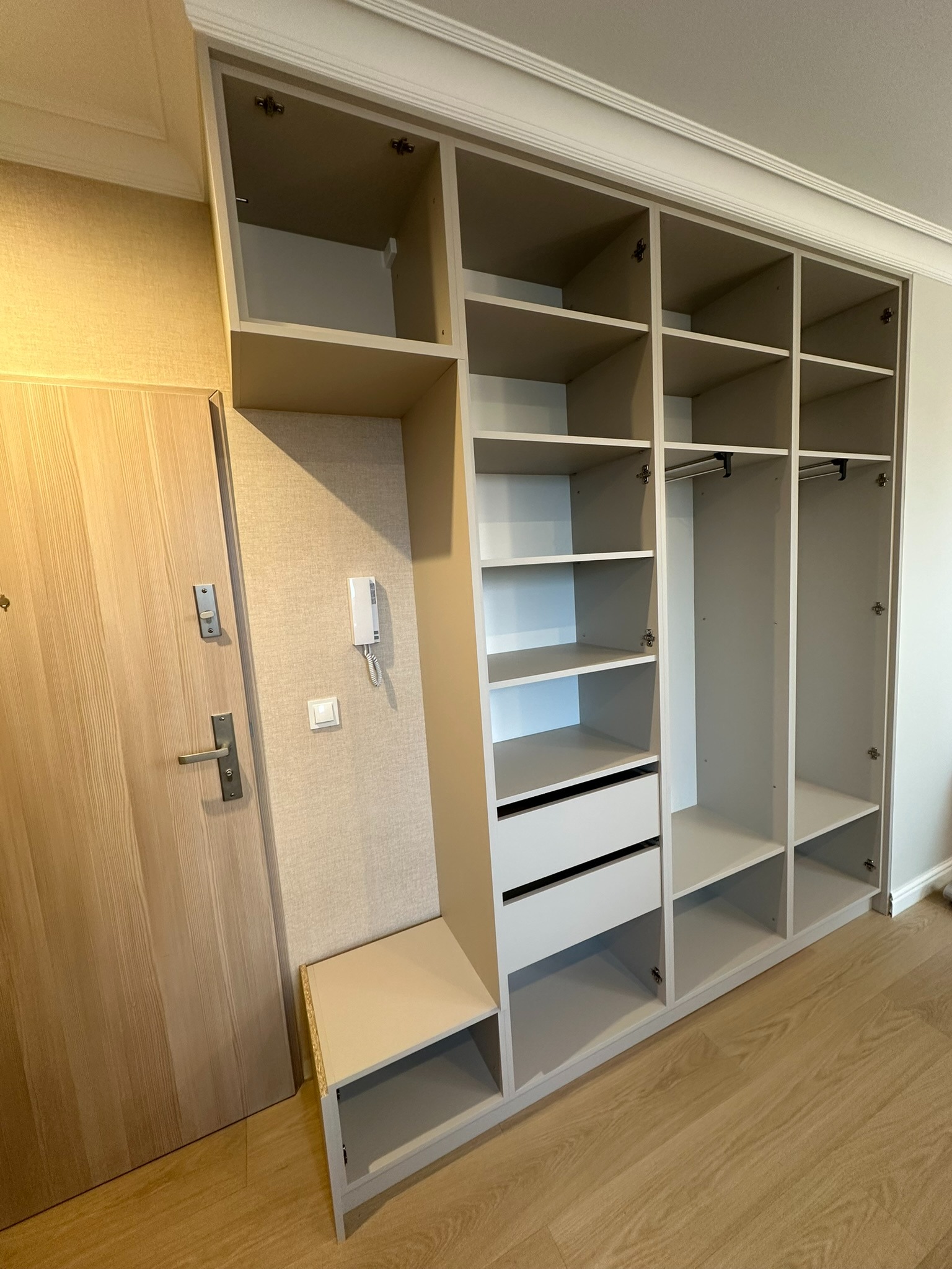 Szafa wnękowa w jasnych odcieniach szarości z półkami, szufladami i drążkami na ubrania, widoczna obok drzwi wejściowych i domofonu.