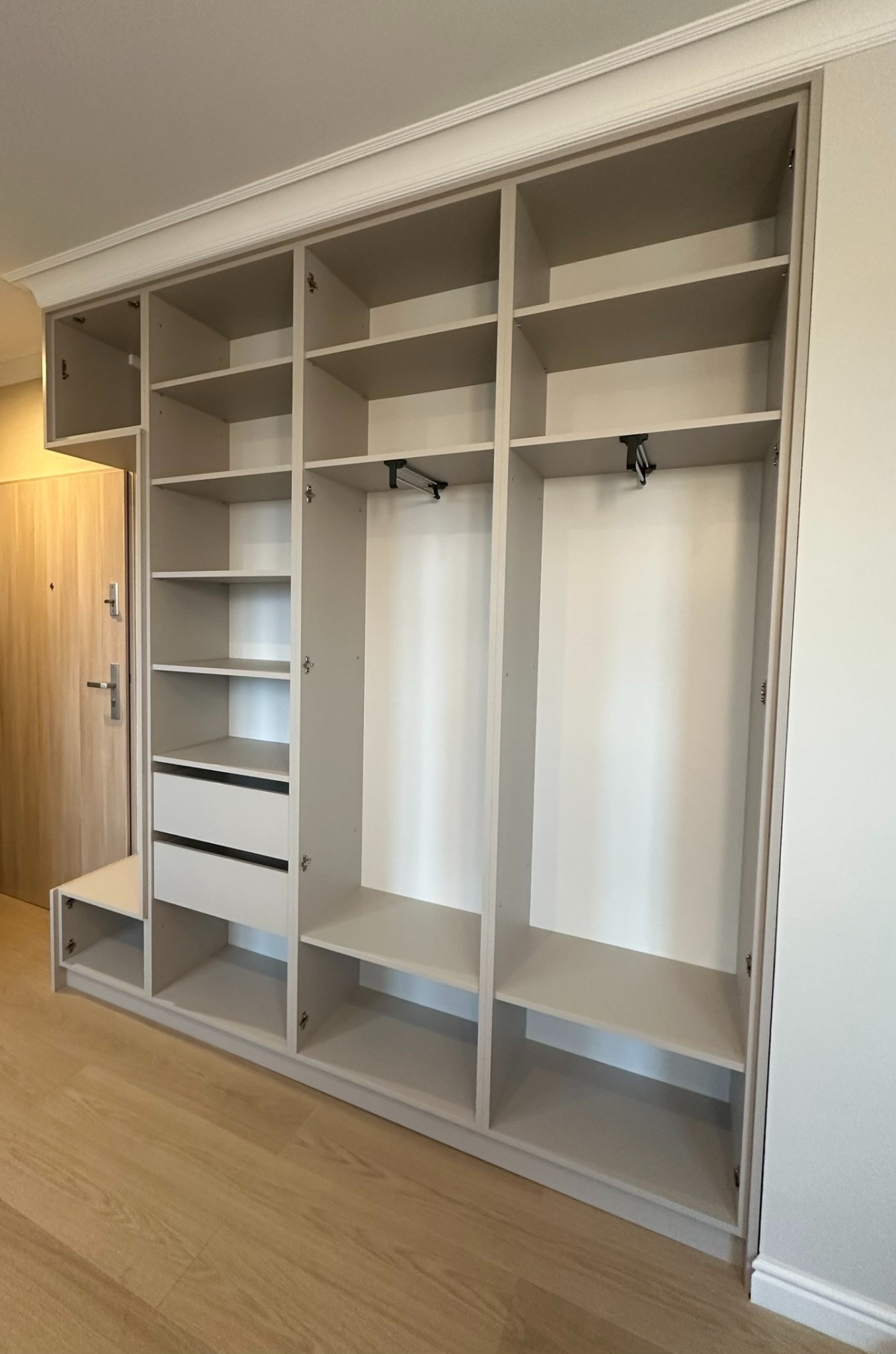 Jasna, zabudowana szafa z półkami i szufladami, widoczne drążki na ubrania, zamontowana w przedpokoju obok drzwi wejściowych.