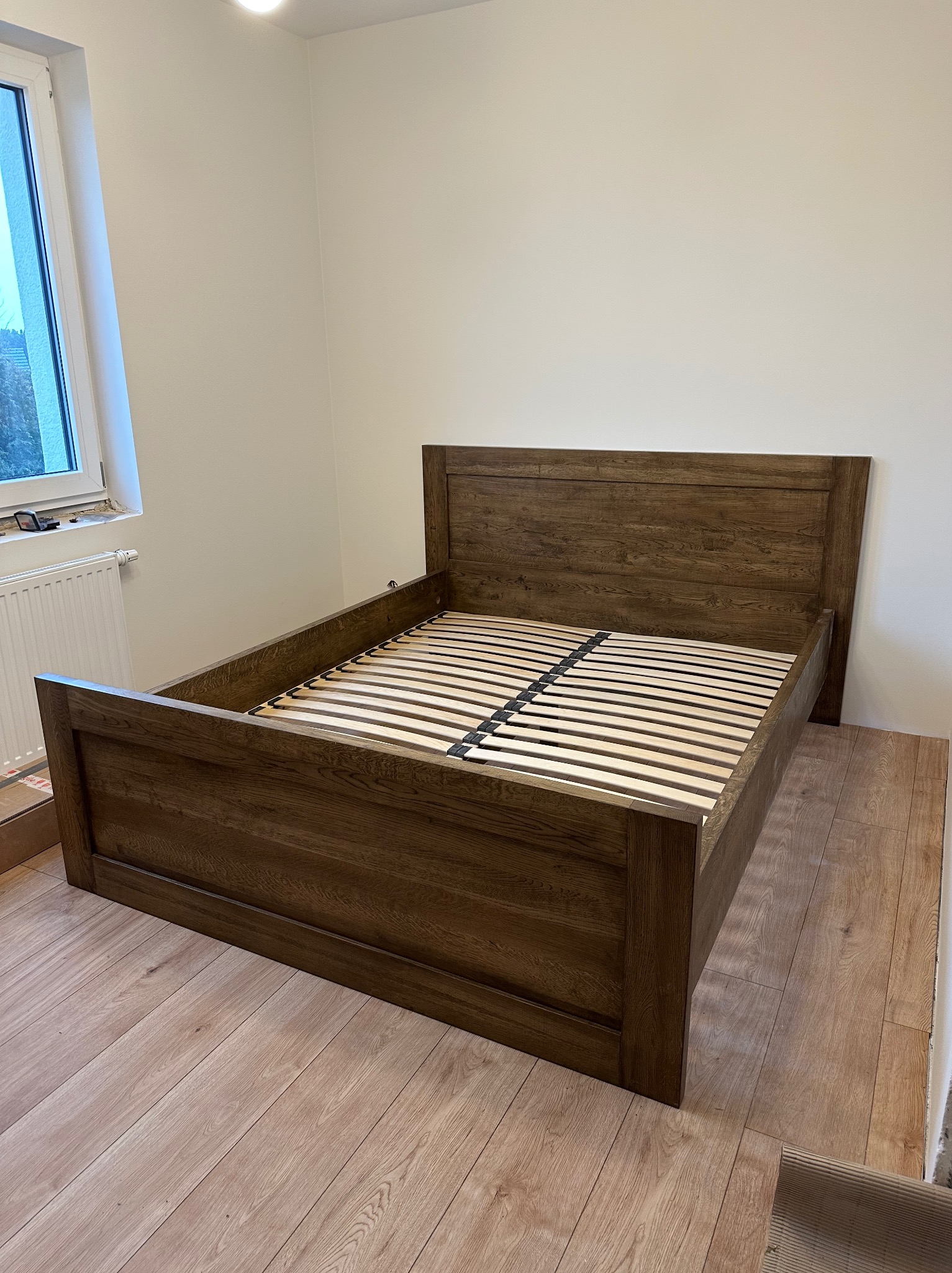 Drewniane łóżko z listwowym stelażem, ustawione na panelach podłogowych, widok z przodu, neutralne oświetlenie.