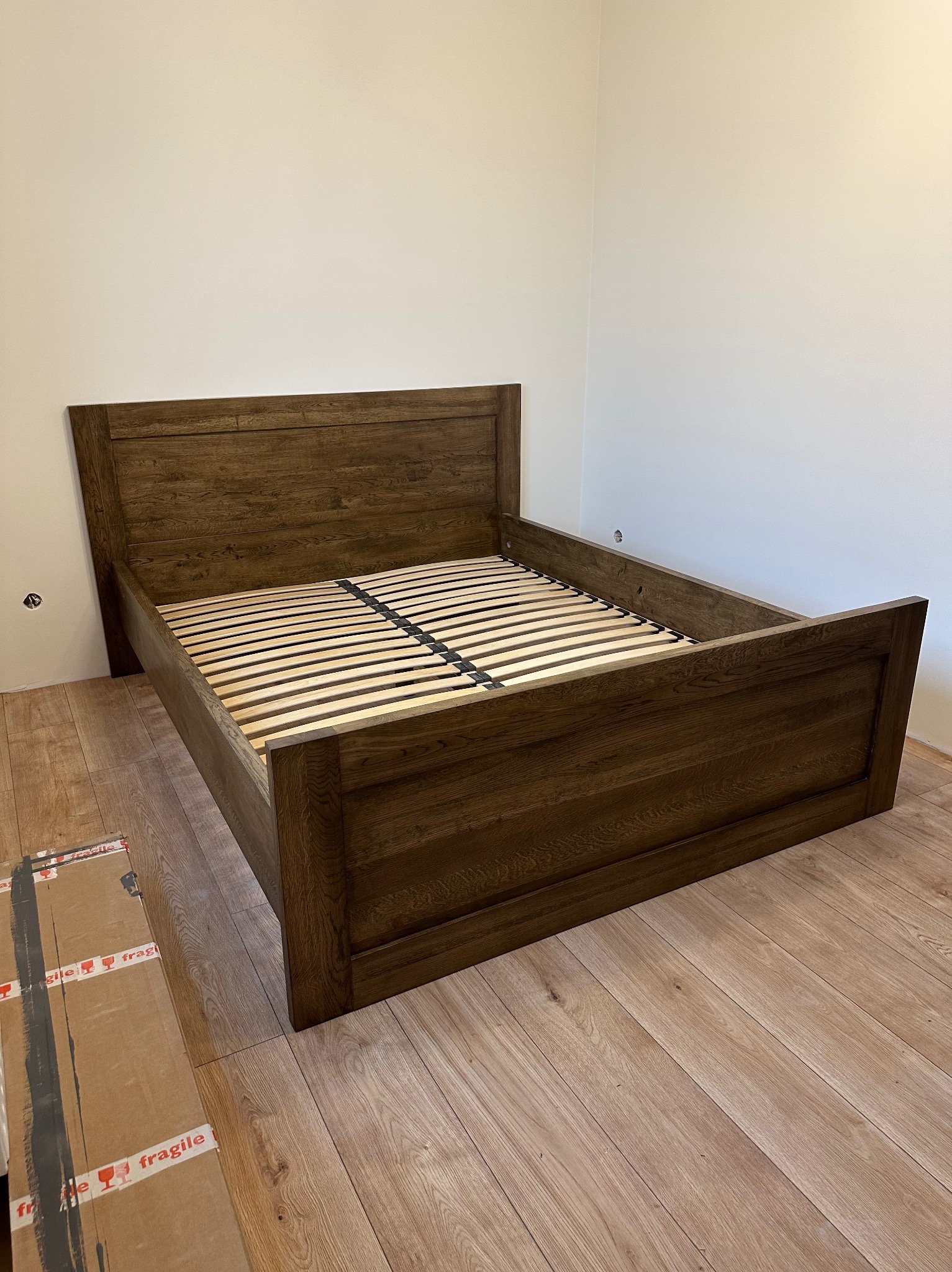Drewniane łóżko z ciemnego drewna z zagłówkiem i drewnianym stelażem, częściowo rozpakowane, karton z napisem 'fragile' obok