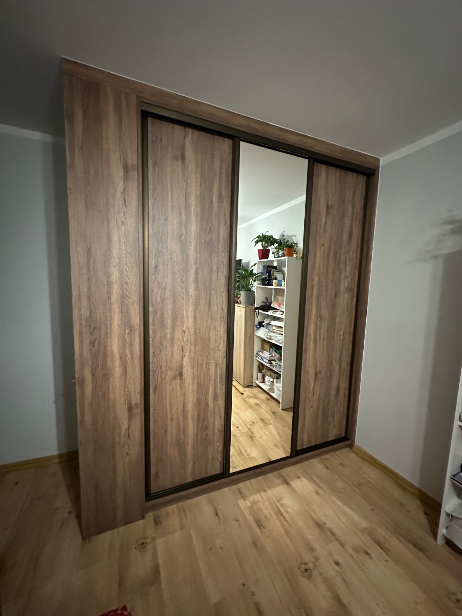 Wysoka szafa przesuwna z lustrzanym frontem, wykończona okleiną w odcieniach brązu, z widocznym odbiciem wnętrza pokoju i regału z roślinami.