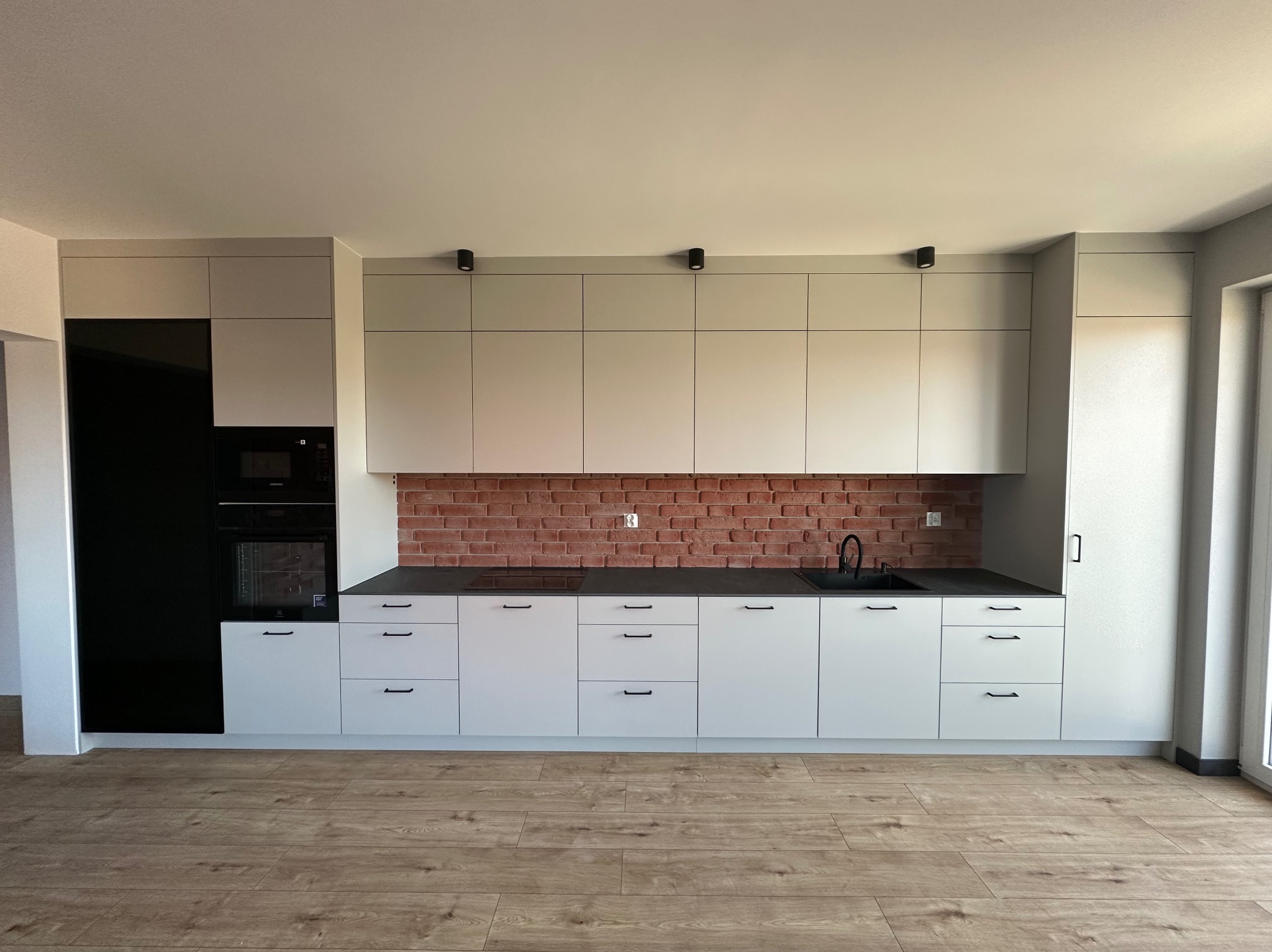 Jasnoszara zabudowa kuchenna z czarnymi elementami AGD i blatem, ceglana ściana nad blatem, drewniana podłoga, minimalistyczne uchwyty.