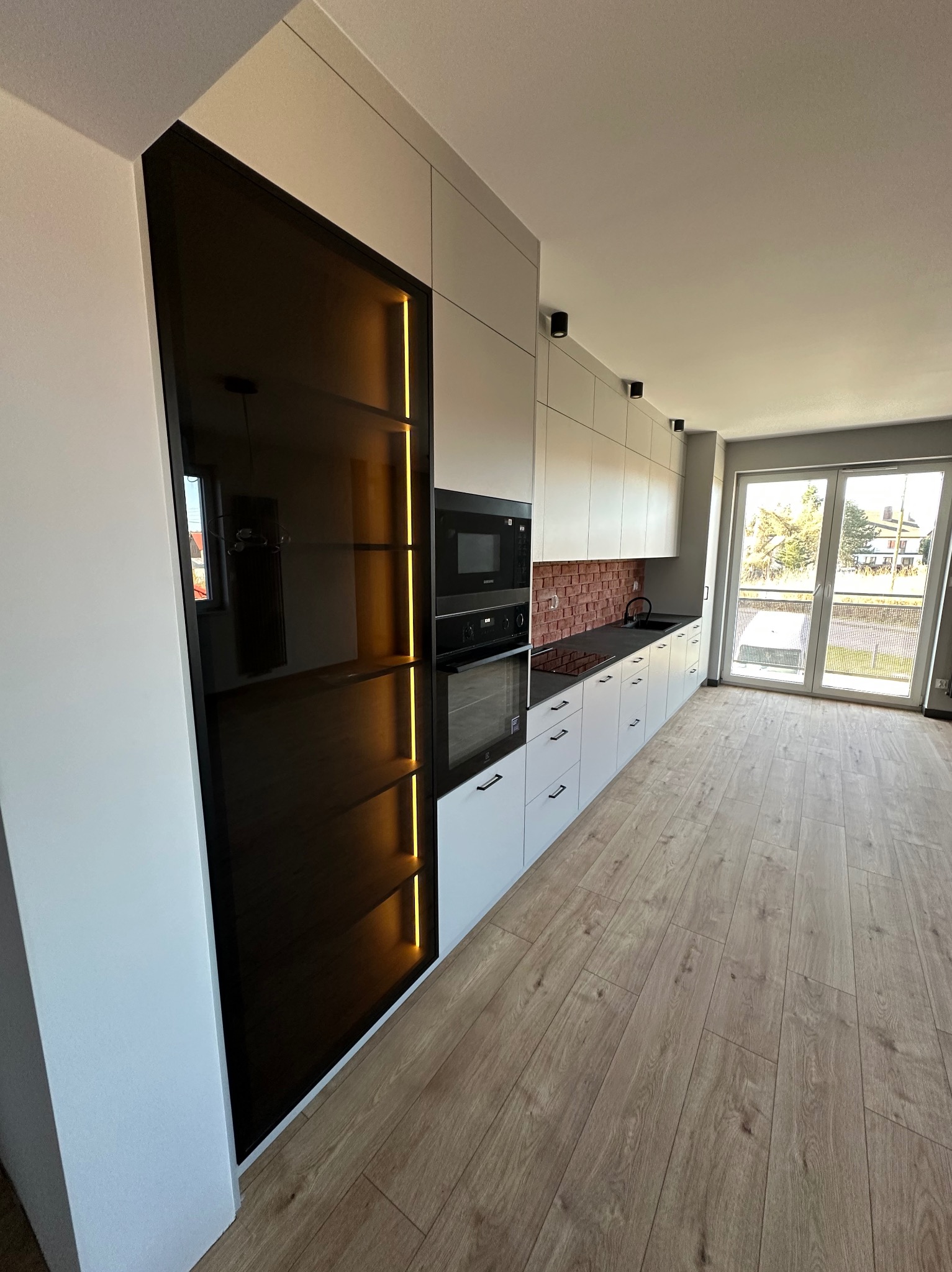 Nowoczesna kuchnia z zabudową w kolorze złamanej bieli, czarnym piekarnikiem i mikrofalówką, oraz szafką z czarnym szkłem podświetloną pionowymi paskami LED, wykończona czerwoną cegłą nad blatem...