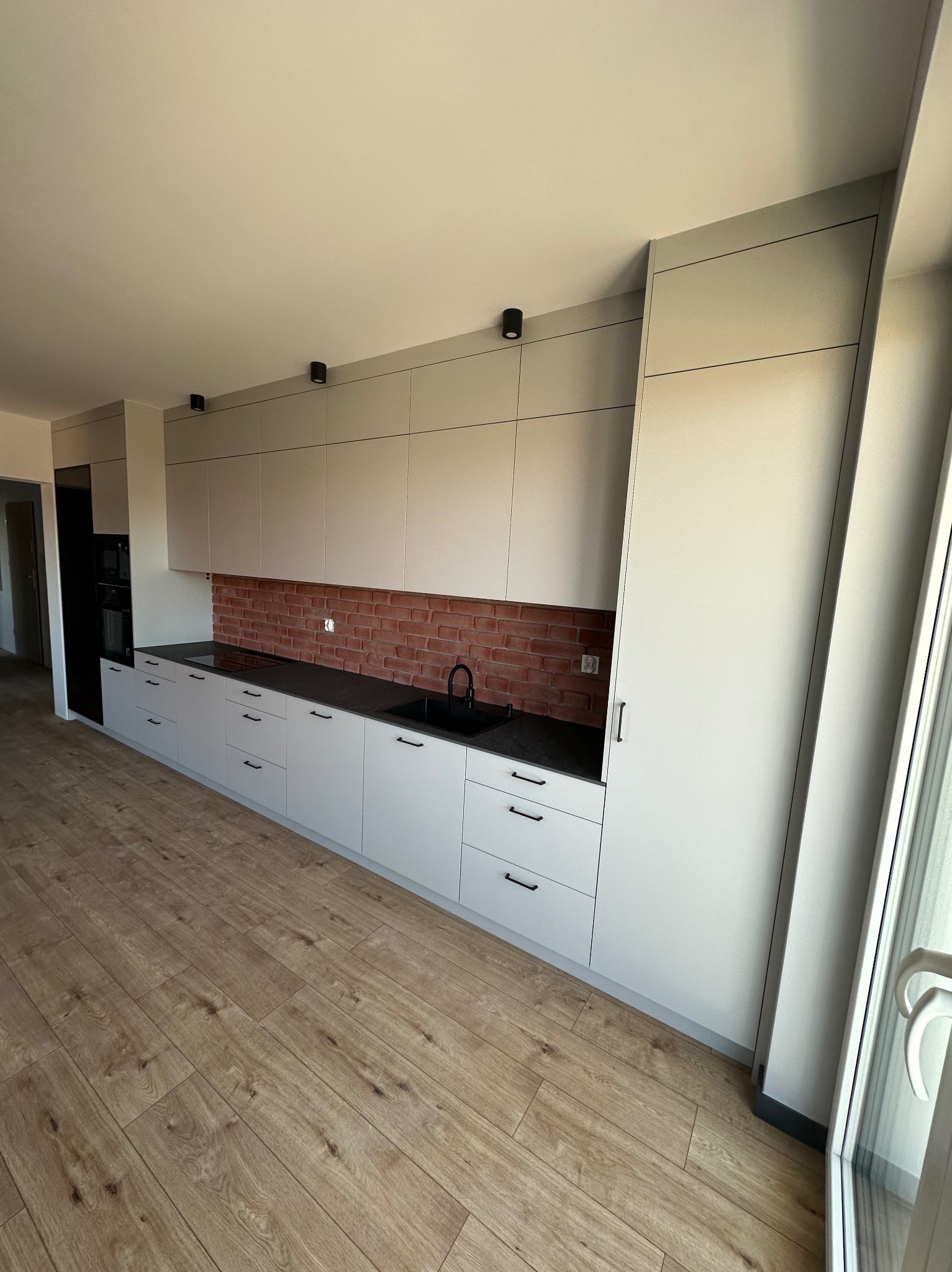 Jasnoszara zabudowa kuchenna z czarnym blatem i baterią, ceglana ściana nad blatem, oświetlenie punktowe sufitowe, drewniana podłoga.