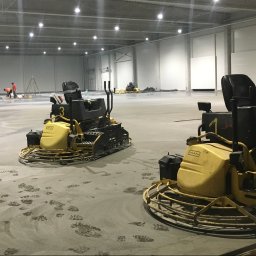 SDI - Dwie zacieraczki mechaniczne do betonu na świeżo wylanej posadzce przemysłowej w hali. Widoczne ślady stóp i pracownicy w oddali.