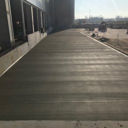 SDI - Świeżo wylana, gładka wylewka betonowa na terenie przemysłowym, z widocznymi pracownikami i maszynami w tle. Jasne światło podkreśla strukturę betonu.