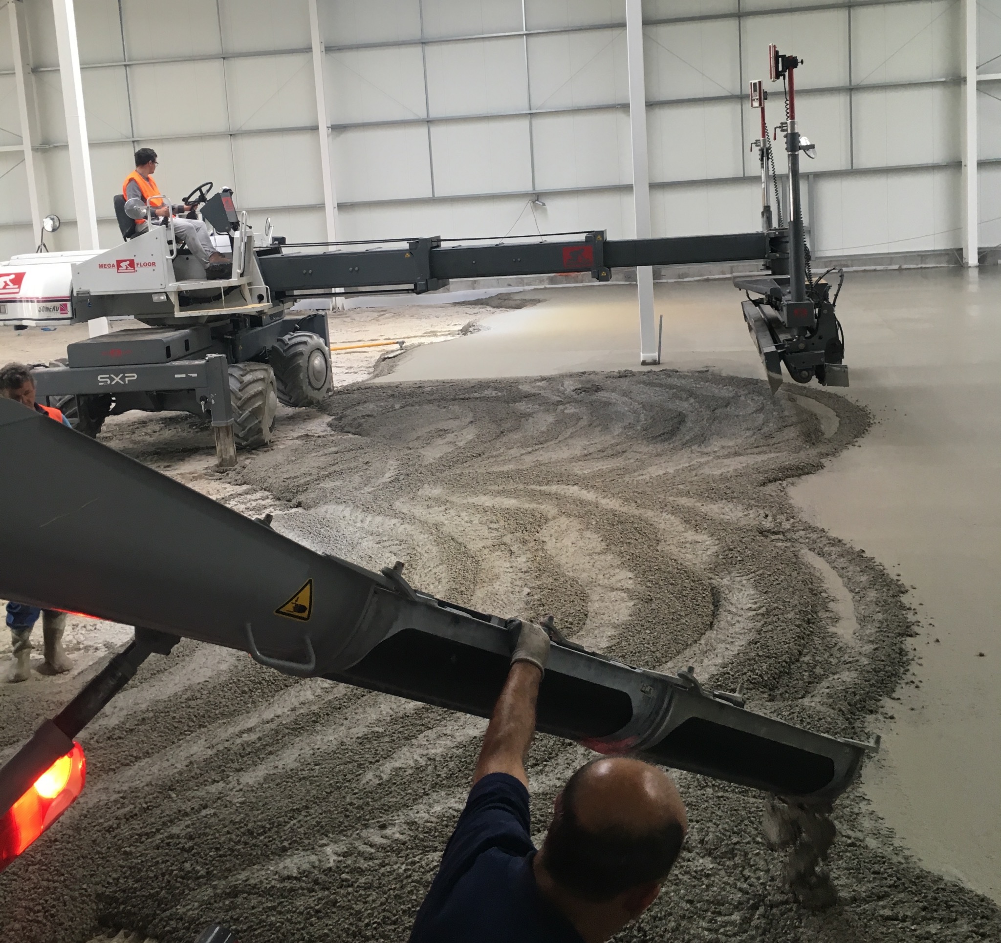 Wylewka posadzki przemysłowej w hali: operator na maszynie Mega Floor SXP rozprowadza beton, drugi pracownik kontroluje proces z bliska. Widoczne świeże i gładkie fragmenty.