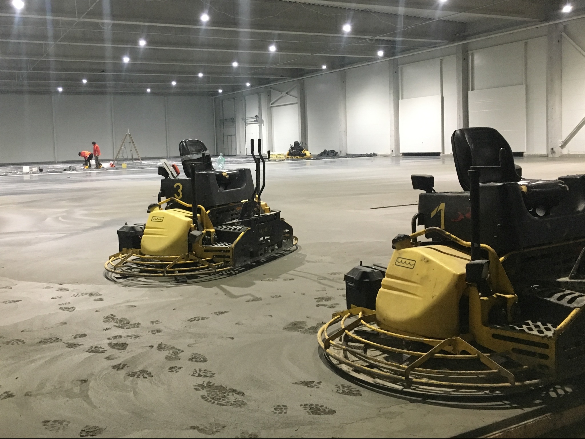 Dwie zacieraczki mechaniczne do betonu na świeżo wylanej posadzce przemysłowej w hali. Widoczne ślady stóp i pracownicy w oddali.