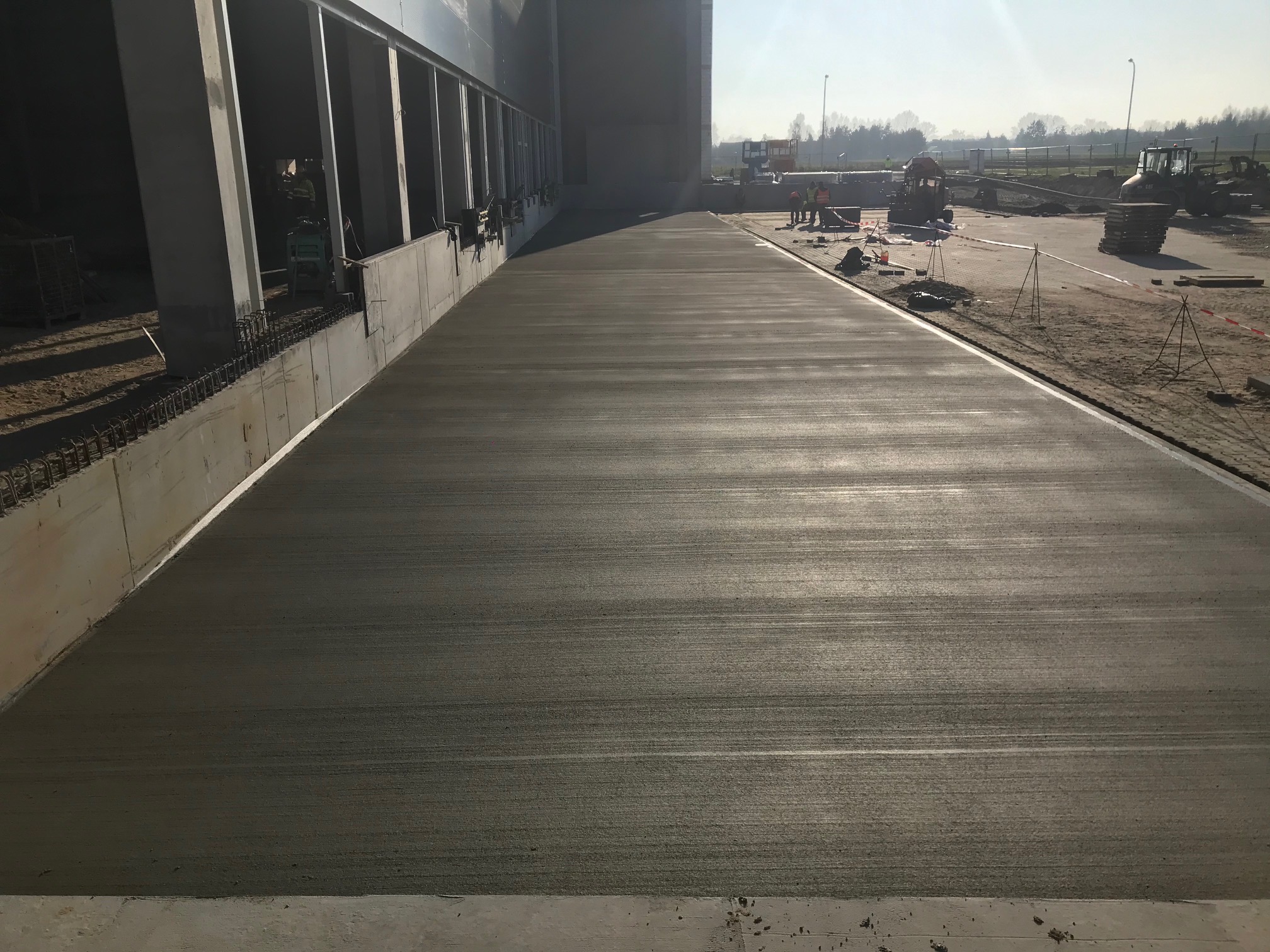 Świeżo wylana, gładka wylewka betonowa na terenie przemysłowym, z widocznymi pracownikami i maszynami w tle. Jasne światło podkreśla strukturę betonu.